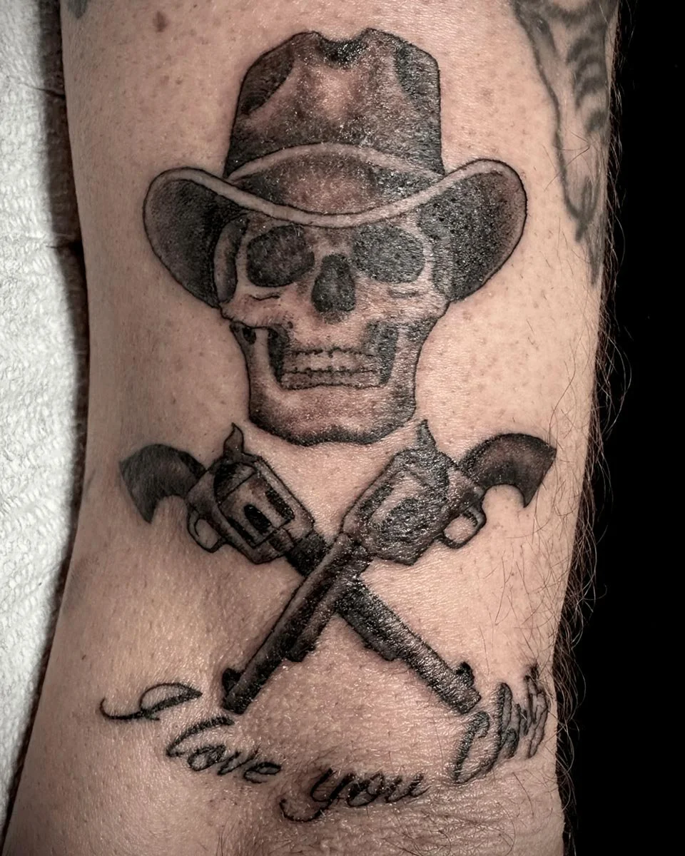 Black and grey skull with a cowboy hat and lettering tattoo on arm.