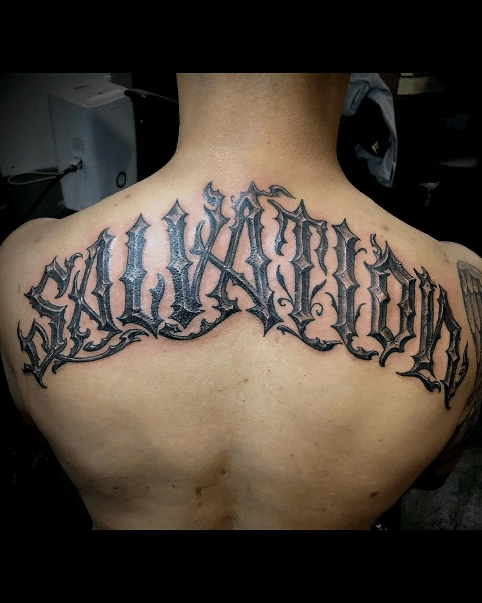 Salvation script tattoo from shoulder to shoulder tattooed in black and grey and fineline style.