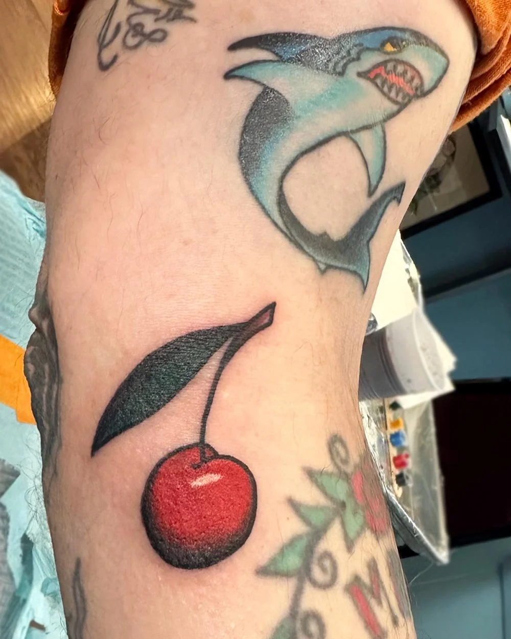 Healed blue shark tattoo next to a fresh cherry color tattoo by John Reardon on an arm.