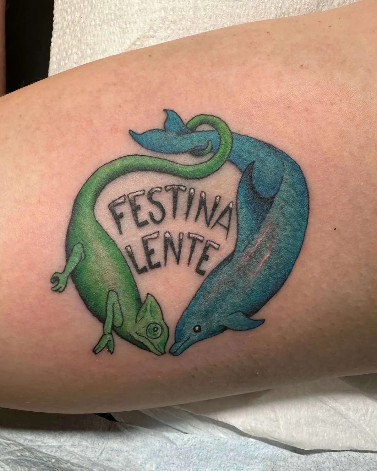 Script tattoo with green and blue lizards holding tails.