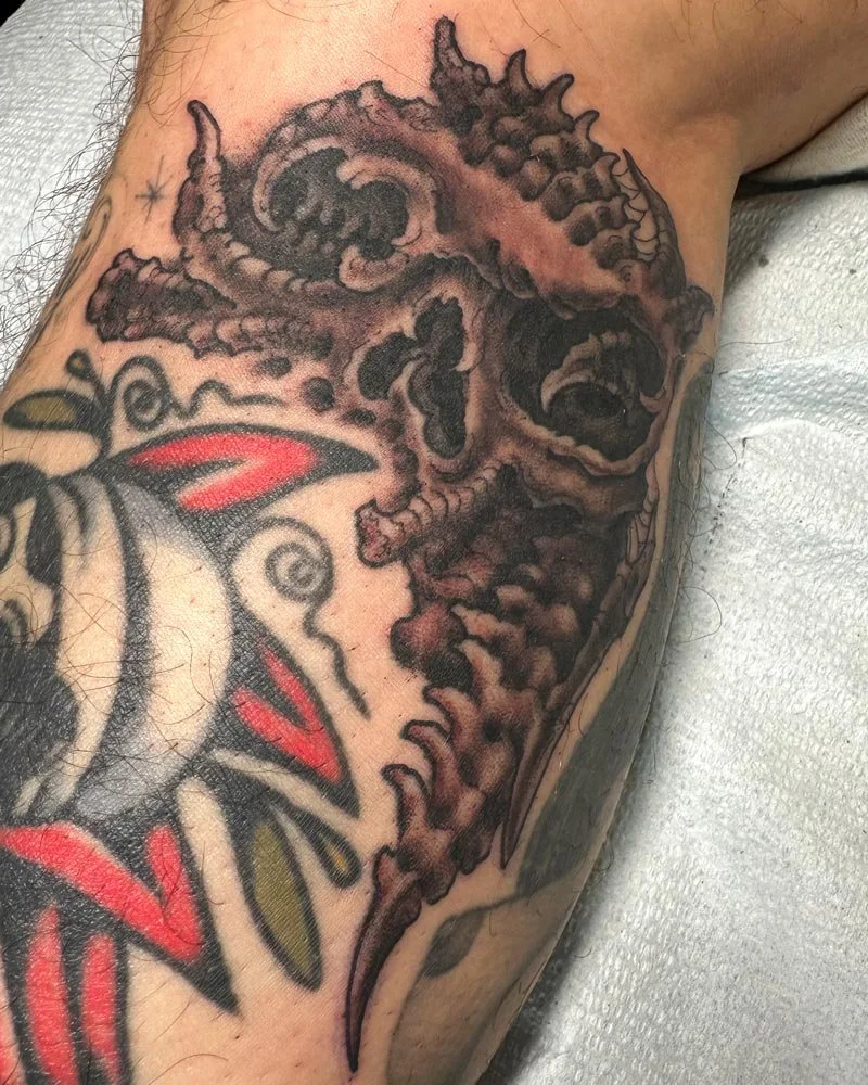 Drawn-on tattoo of a black and grey biomech skull above a healed color tattoo.