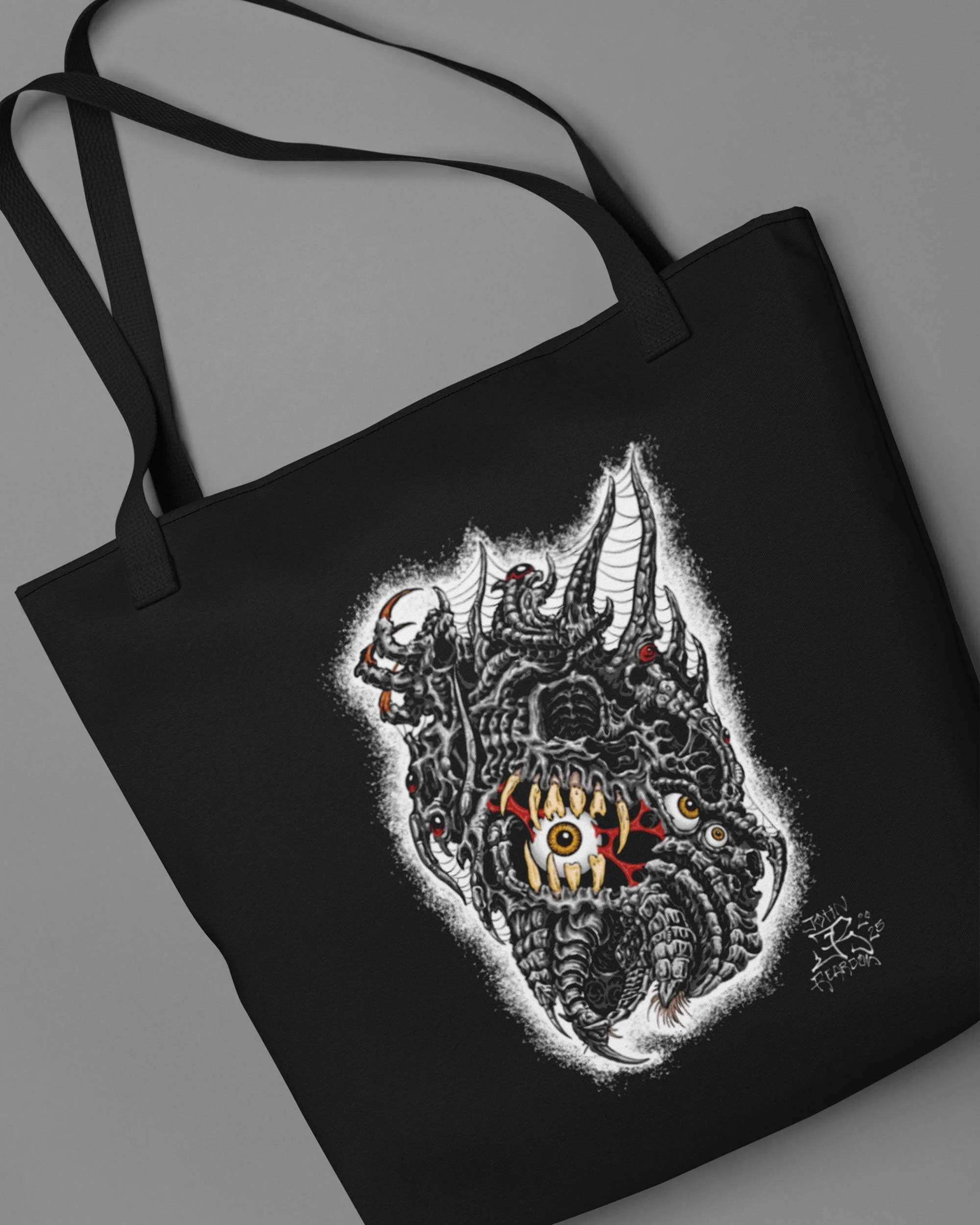 Black tote bag laying down on a grey background with-tattooer-John Reardon artwork of biomech creature with eyeballs.