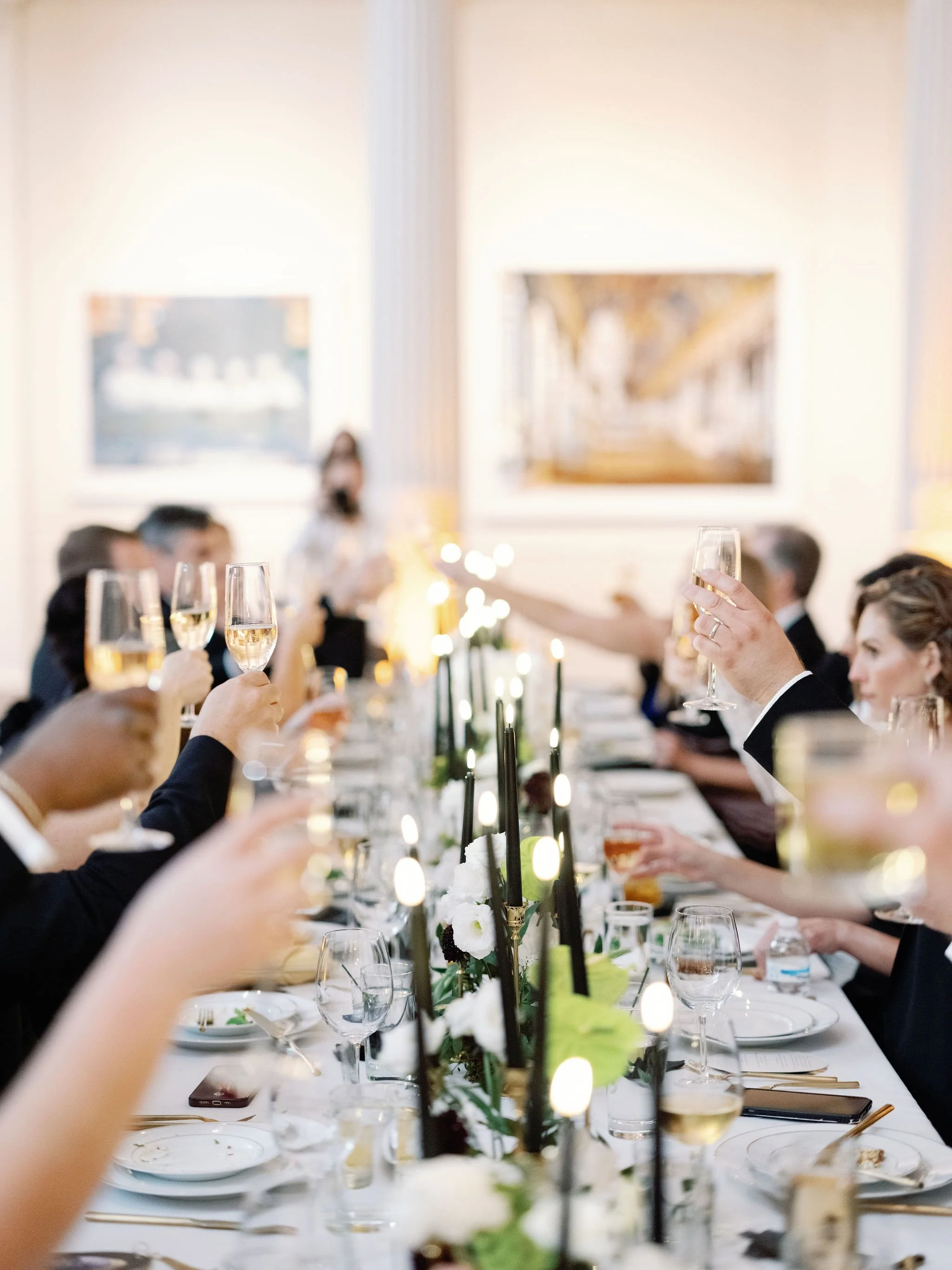 People seated at a long elegant table celebrating with toast in a formal dining setting, with floral centerpiece and candles.
