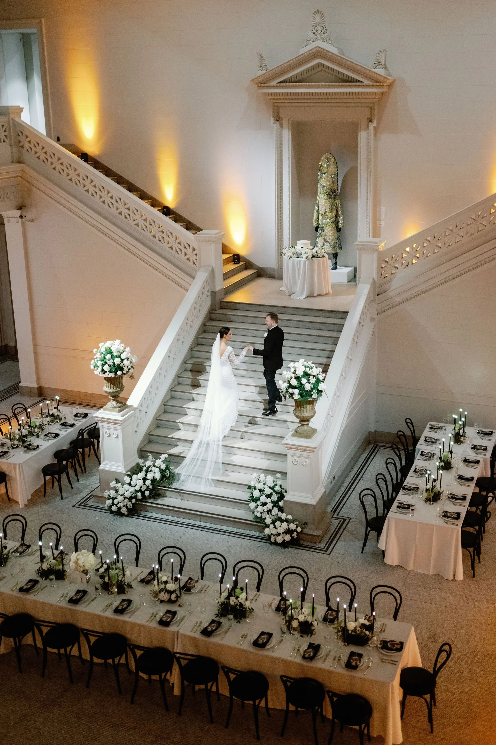 A wedding ceremony taking place in a grand staircase hall with a bride and groom holding hands at the bottom of the stairs, floral arrangements, and elegant table settings for a reception.