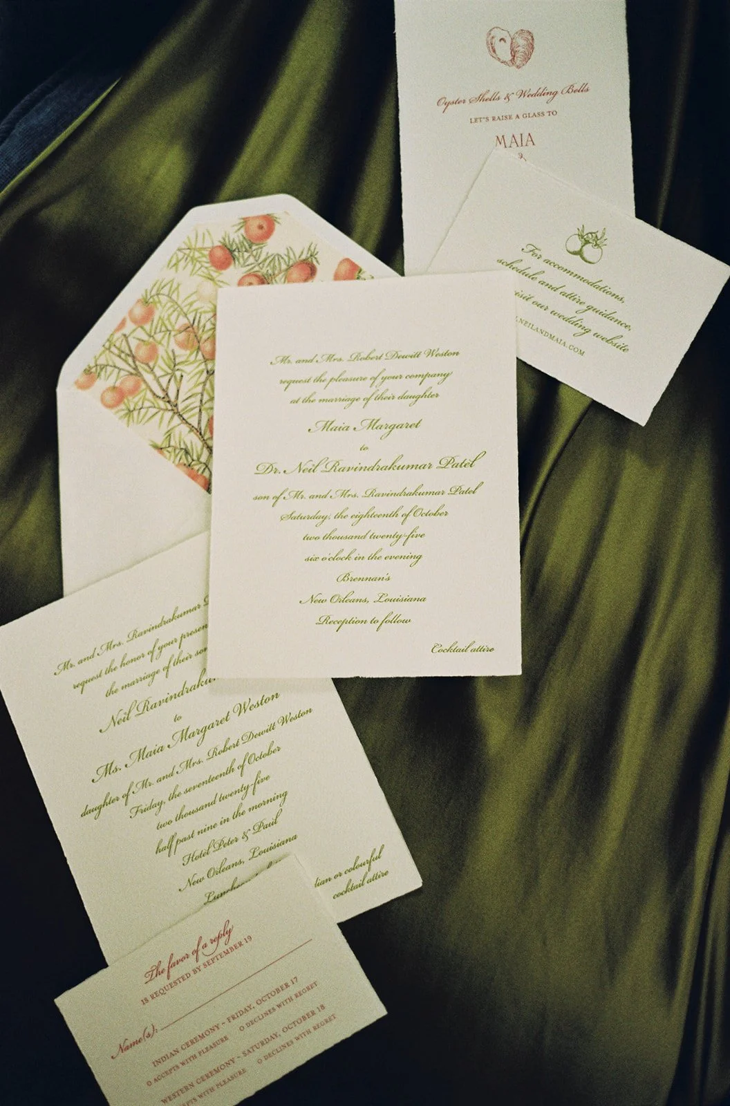 Wedding invitation suite with multiple cards, including a main invitation, RSVP card, and information card, all placed on a dark green fabric with a floral-lined envelope.