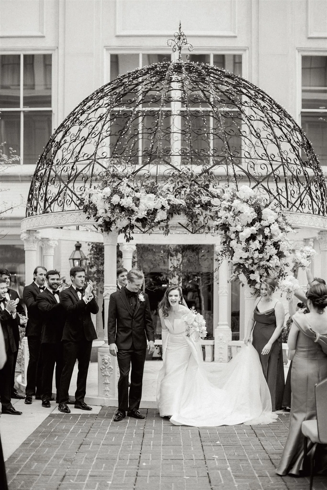 A wedding celebration outside with the bride and groom walking under a decorative iron gazebo adorned with flowers and lights while guests applaud.