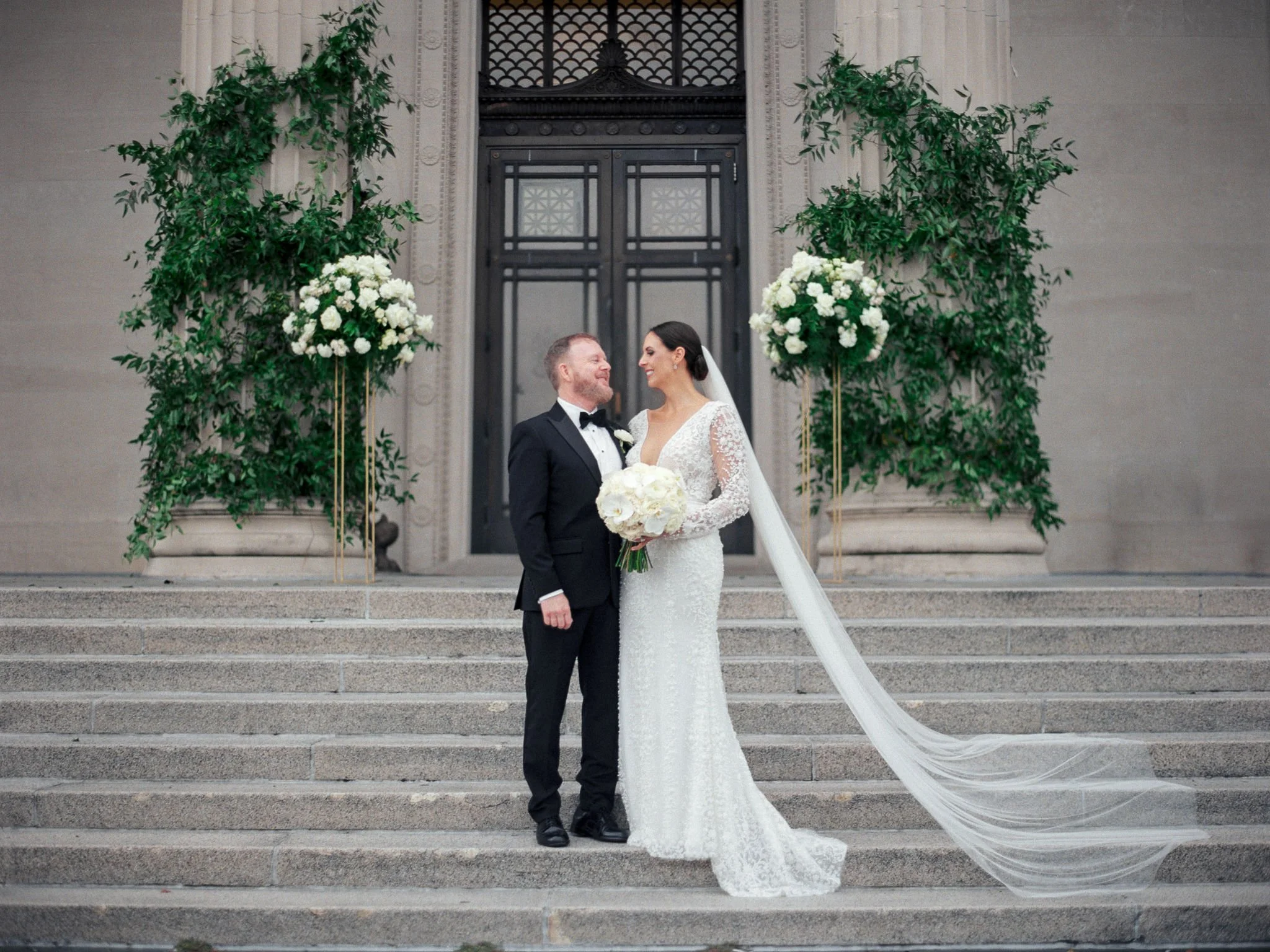 A bride and groom standing on an outdoor staircase, smiling at each other, with large floral arrangements and greenery on either side of them.