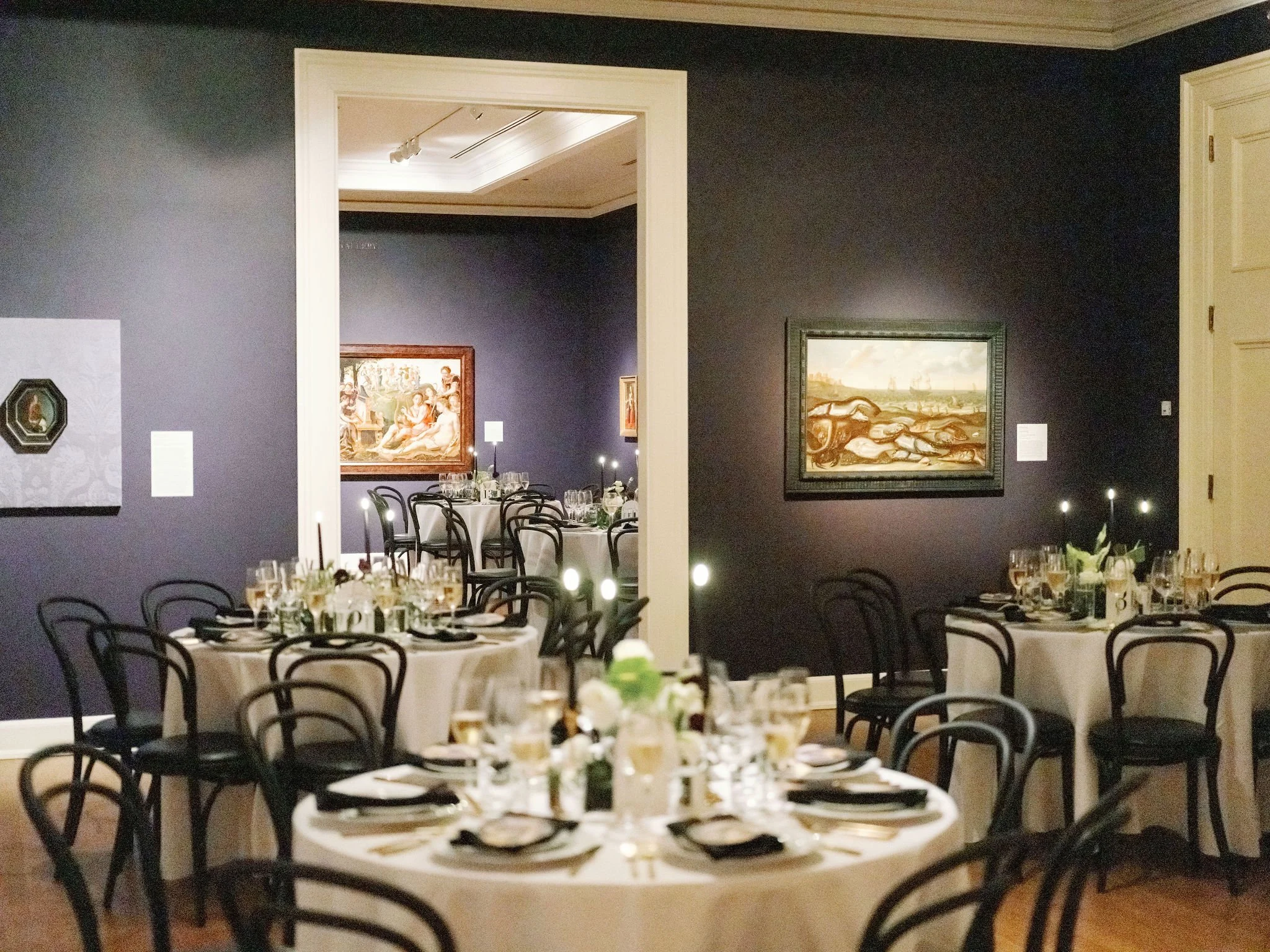 Dining room set up for a formal event with round tables covered with white tablecloths, black chairs, and place settings including plates, utensils, glasses, and floral centerpieces. The walls display framed artwork, and there is a doorway leading to