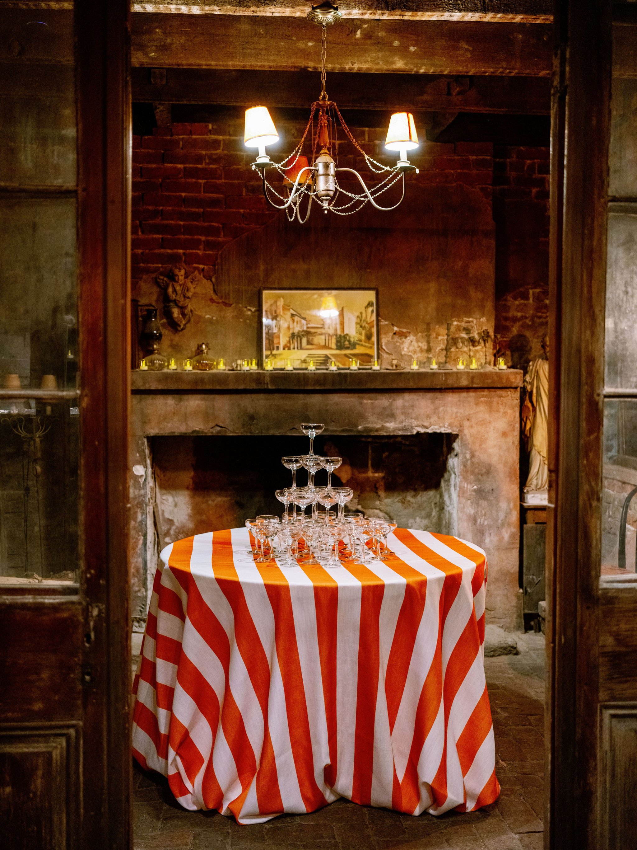 A room with exposed brick walls and wooden beams ceiling, seen through open wooden double doors, with a table covered by a red and white striped tablecloth, on which a pyramid of stacked martini glasses is arranged. A chandelier with five lamps is ha