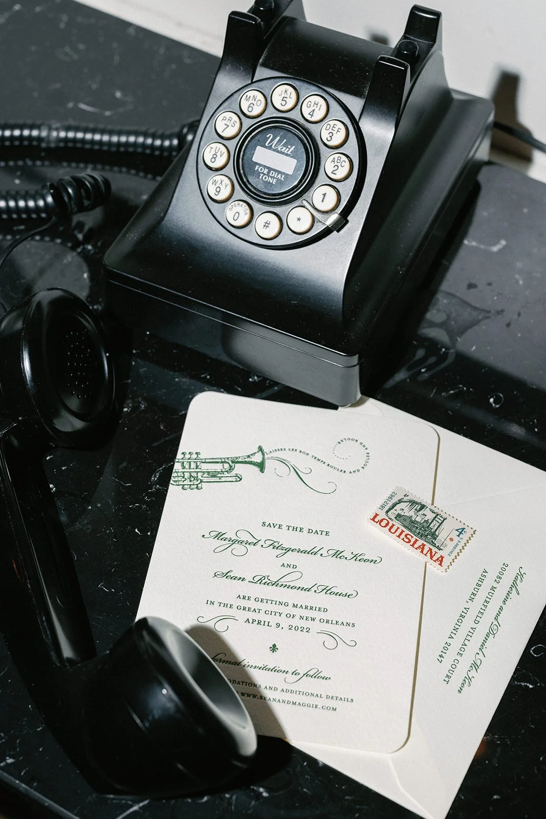A black rotary phone, a black landline phone receiver, and a wedding invitation with a Louisiana stamp on a black surface.