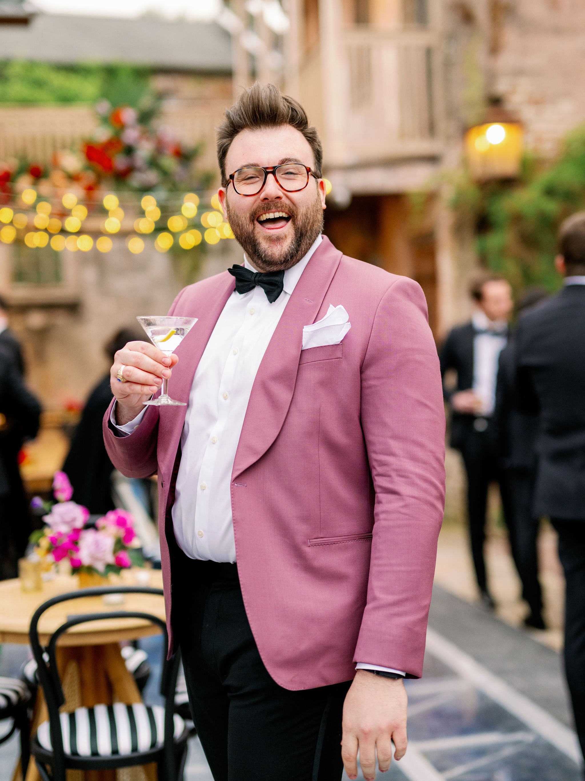 A man with glasses, a beard, and a big smile, wearing a pink tuxedo jacket, white shirt, and black bow tie, is holding a martini glass and enjoying a celebration outdoors.