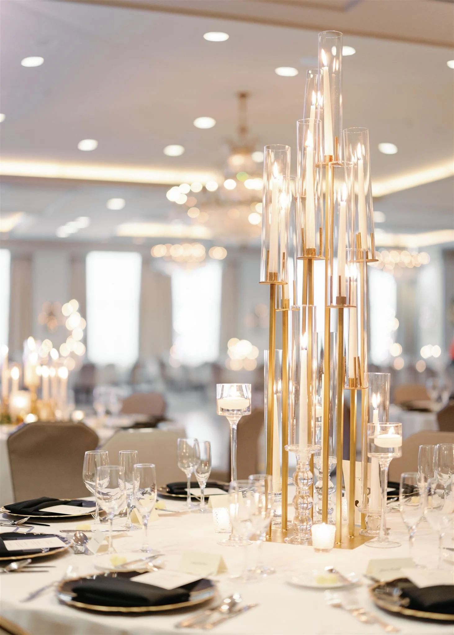 An elegant banquet table setting with wine glasses, black napkins, and a tall centerpiece with candles in glass holders in a well-lit room with a chandelier and large windows.
