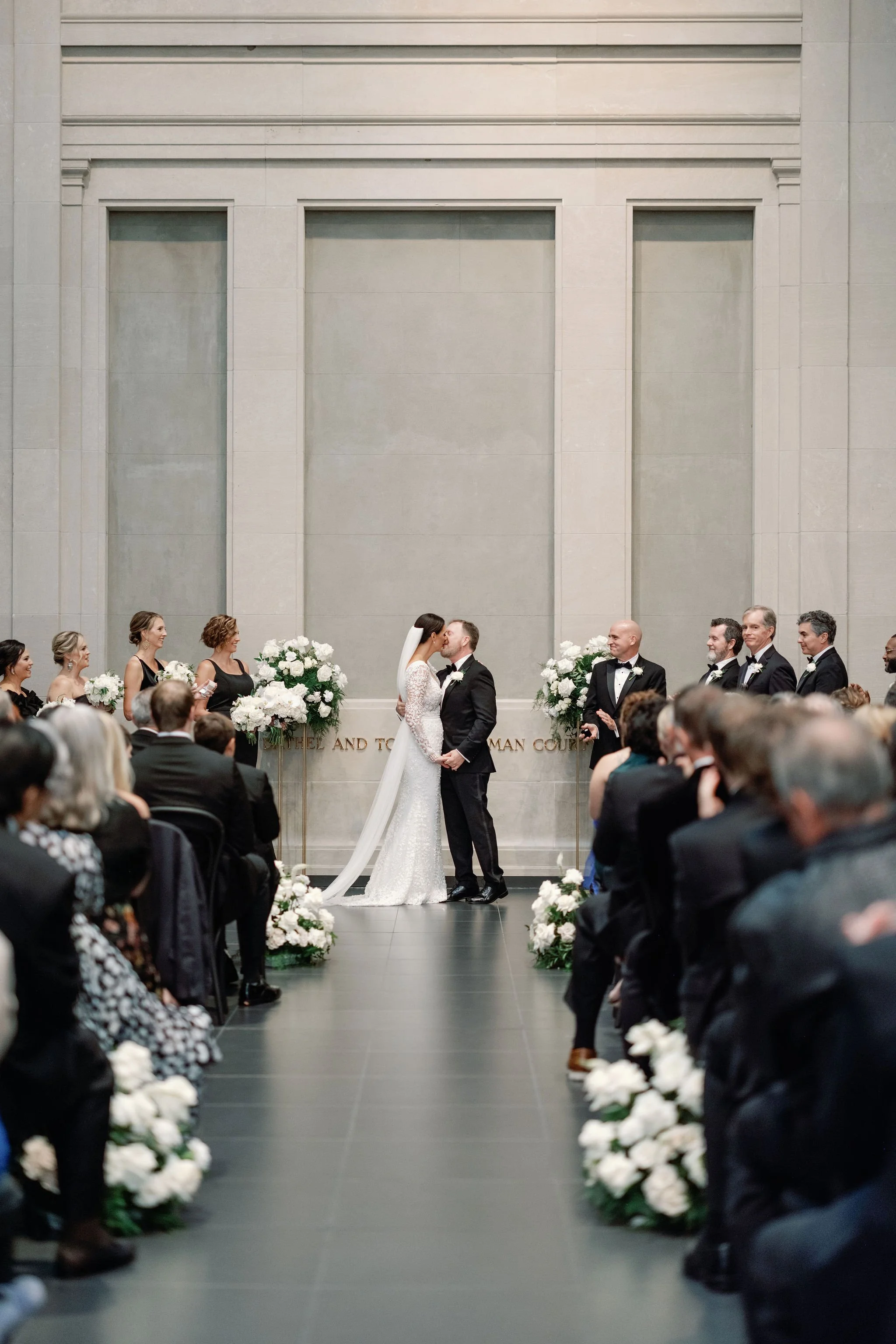 A wedding ceremony inside a large, elegant hall with high walls and minimalist decor. The bride and groom are standing in the center, facing each other, surrounded by bridesmaids and groomsmen. The aisle is decorated with white flowers, and guests ar