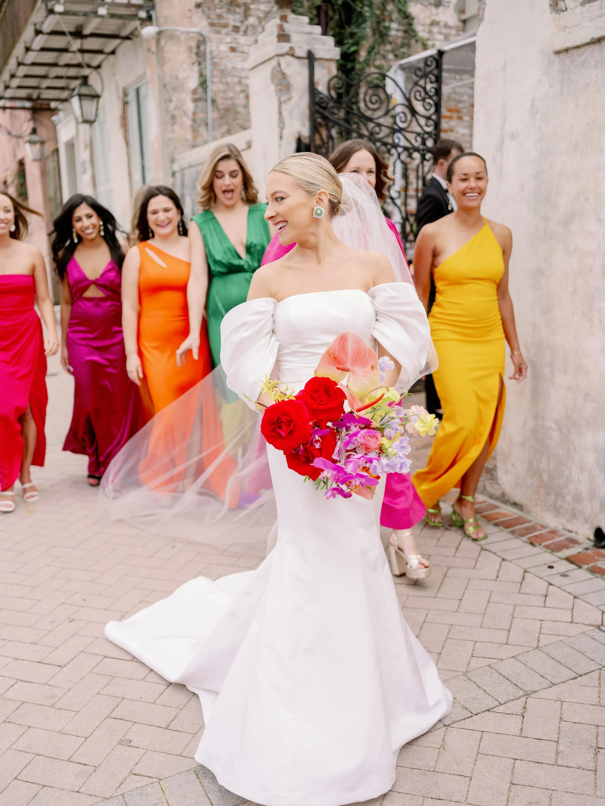 A bride in a white wedding dress holding a colorful bouquet walks with a group of women dressed in vibrant, colorful dresses in an outdoor alleyway with brick and stucco walls.