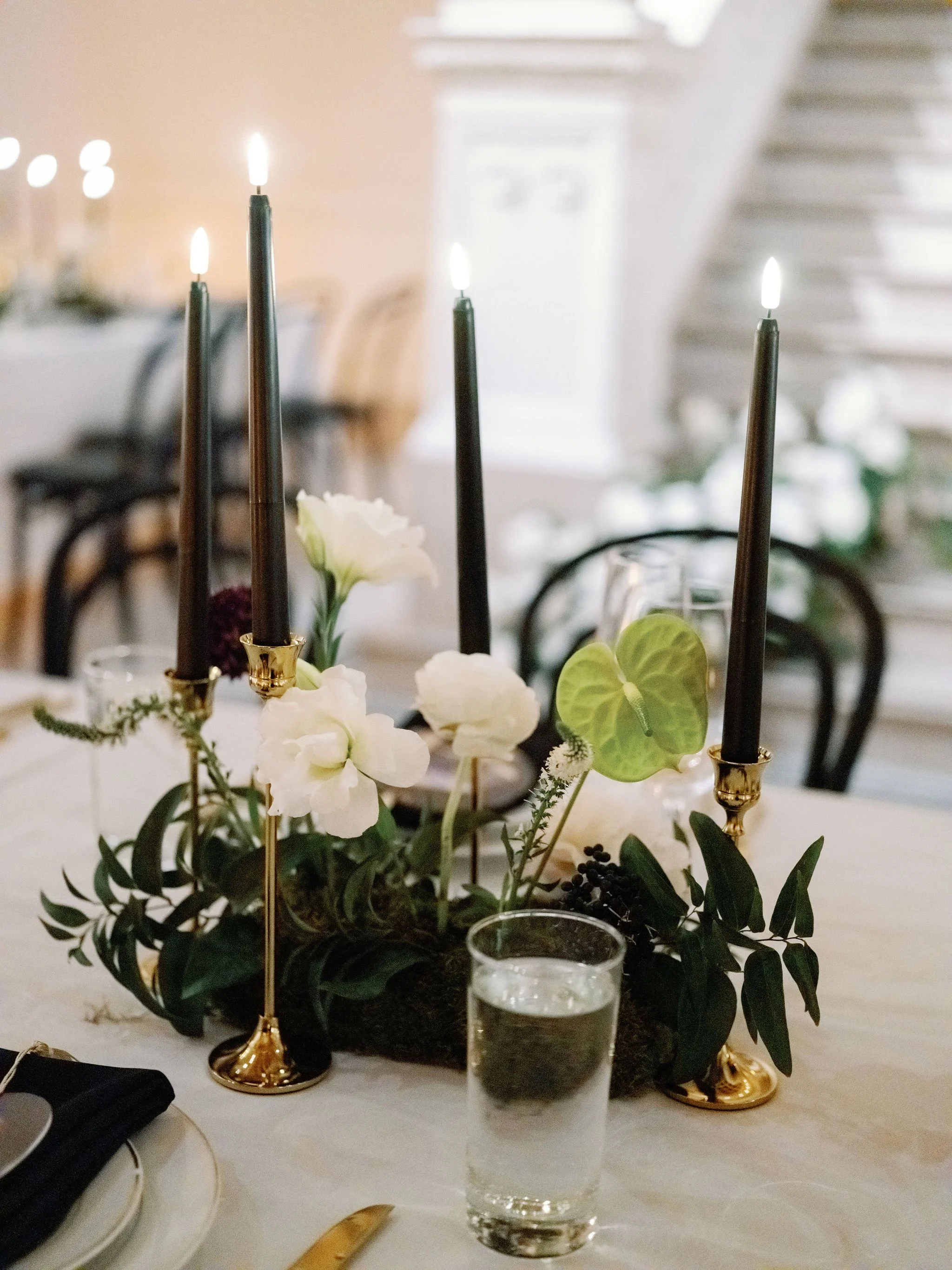 A table centerpiece with white flowers, greenery, and black candles in gold holders at a dining table. A glass of water and a white plate with cutlery are also on the table.