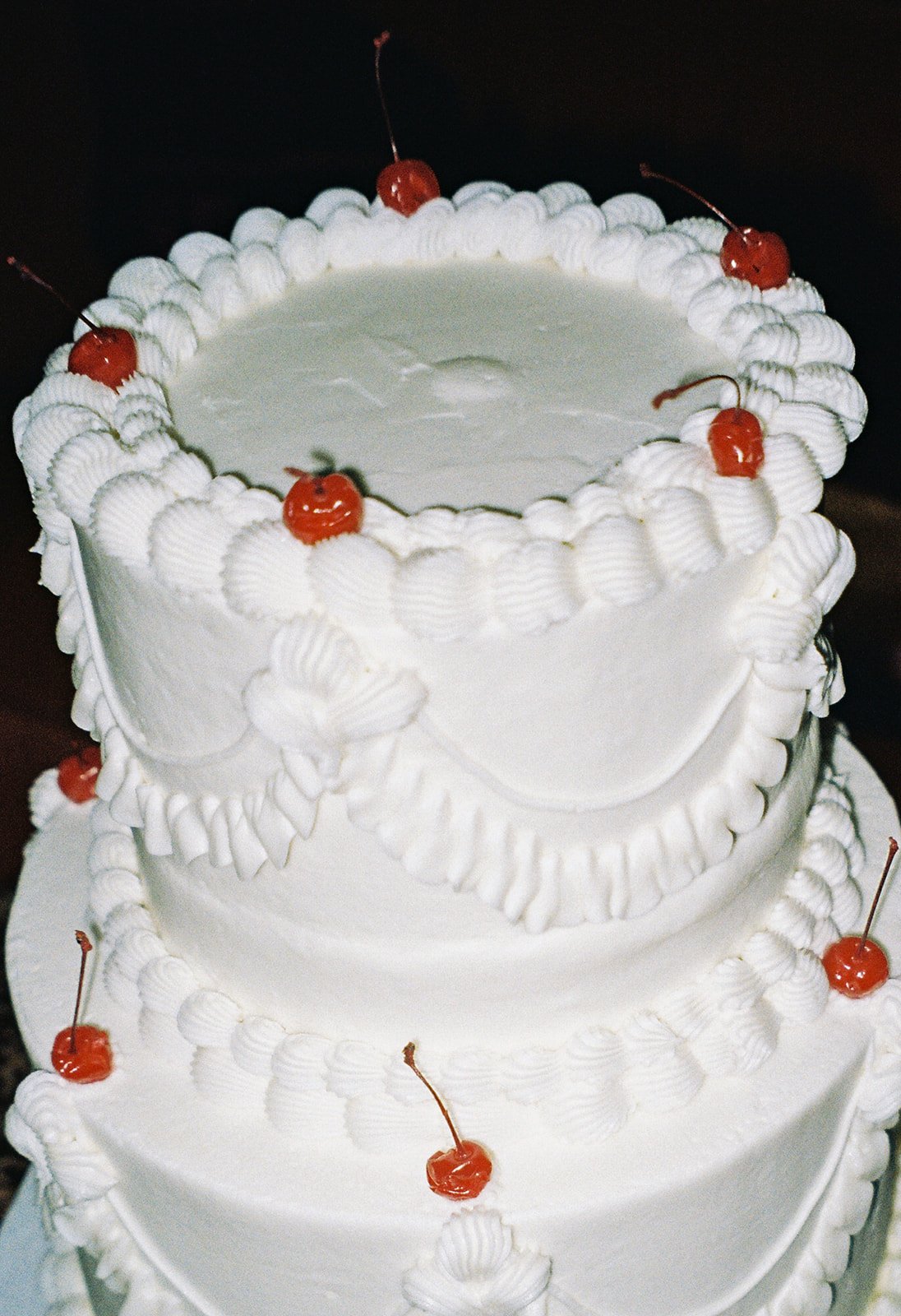 Three-tier vintage white wedding cake decorated with whipped cream and red cherries, with cherries on top and around the edges of each tier.