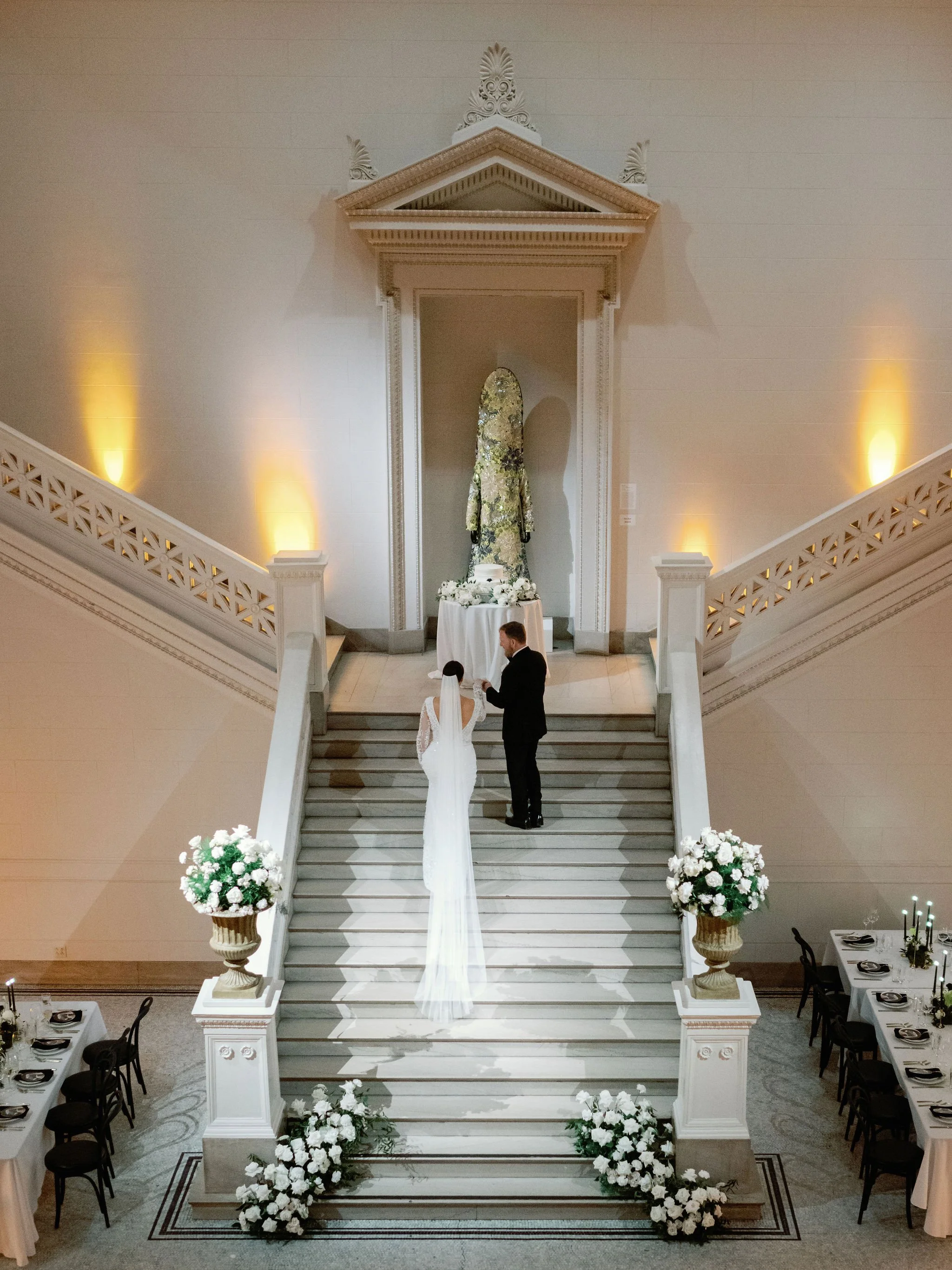 A wedding ceremony taking place at the top of a grand staircase decorated with white flowers in vases on either side. At the top of the stairs, a bride in a white gown and a groom in a black suit are facing each other, holding hands, with an altar fe