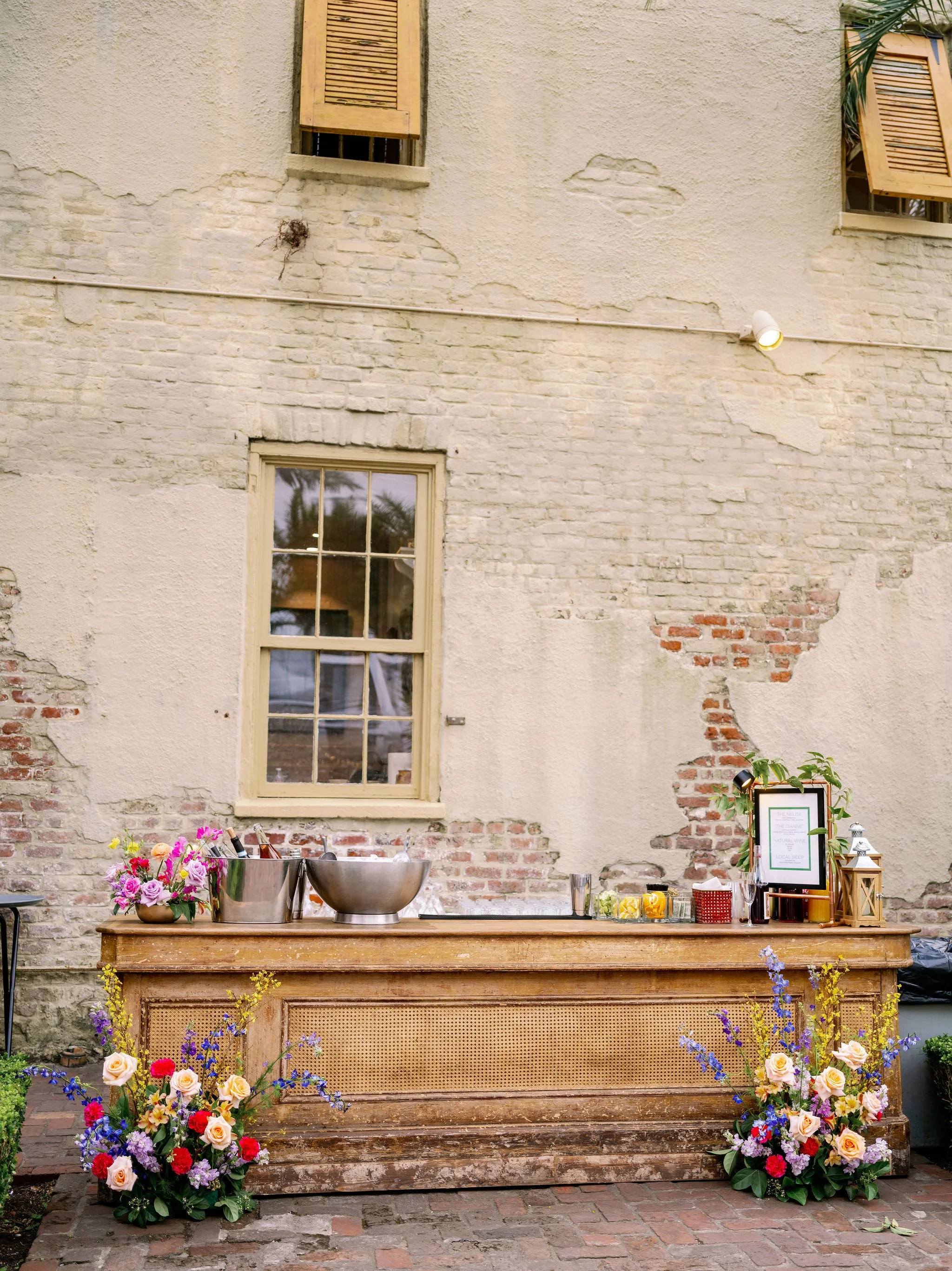 A rustic outdoor bar setup with two large metal bowls, flower arrangements, a framed menu, and various bottles and garnishes, all placed on a wooden table against a weathered brick and plaster wall with windows and greenery.