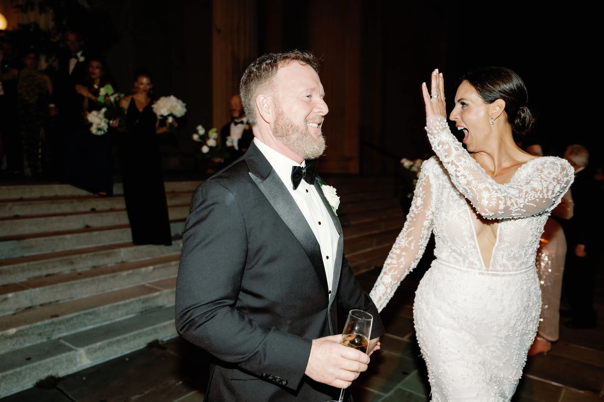 A groom and bride at a wedding reception, smiling and celebrating, with guests in the background holding flowers.