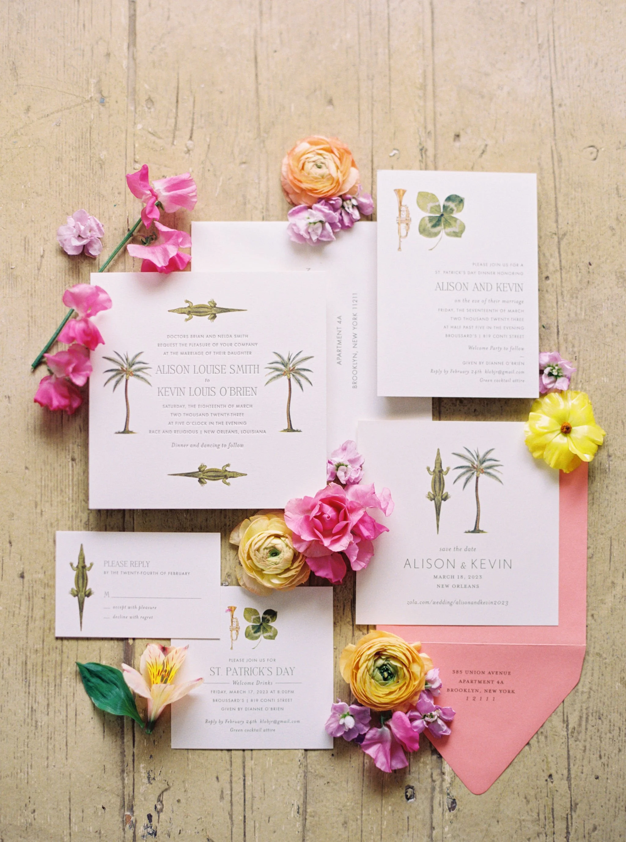 Colorful New Orleans wedding invitations featuring palm tree illustrations, pink, yellow, and peach flowers, scattered on a wooden surface with pink and yellow paper backing.
