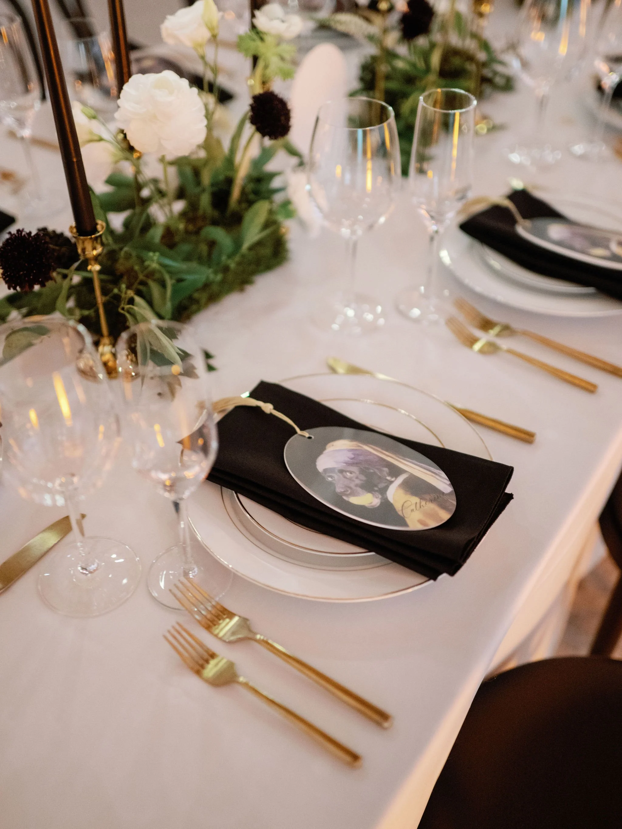 Elegant table setting with gold utensils, black napkins, clear wine glasses, a floral centerpiece with white and dark flowers, and a place card with a portrait picture.