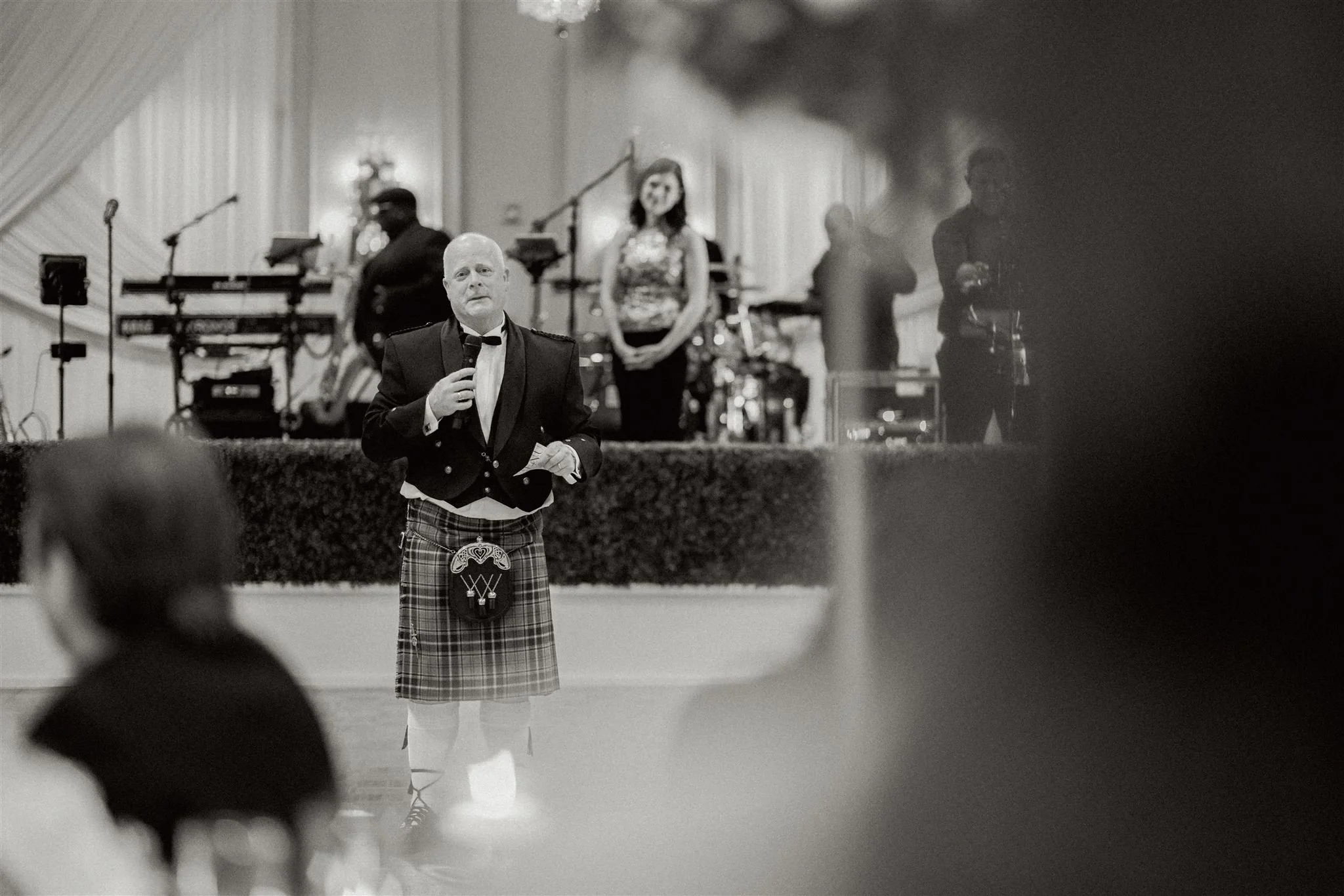 Man dressed in a tuxedo and kilt speaking into a microphone in a ballroom with a band playing in the background.