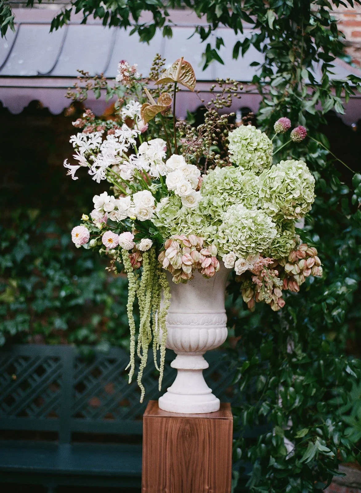 A large white vase filled with various white and light pink flowers, including hydrangeas, roses, and other blooms, sits on a wooden pedestal outdoors, with green foliage in the background.