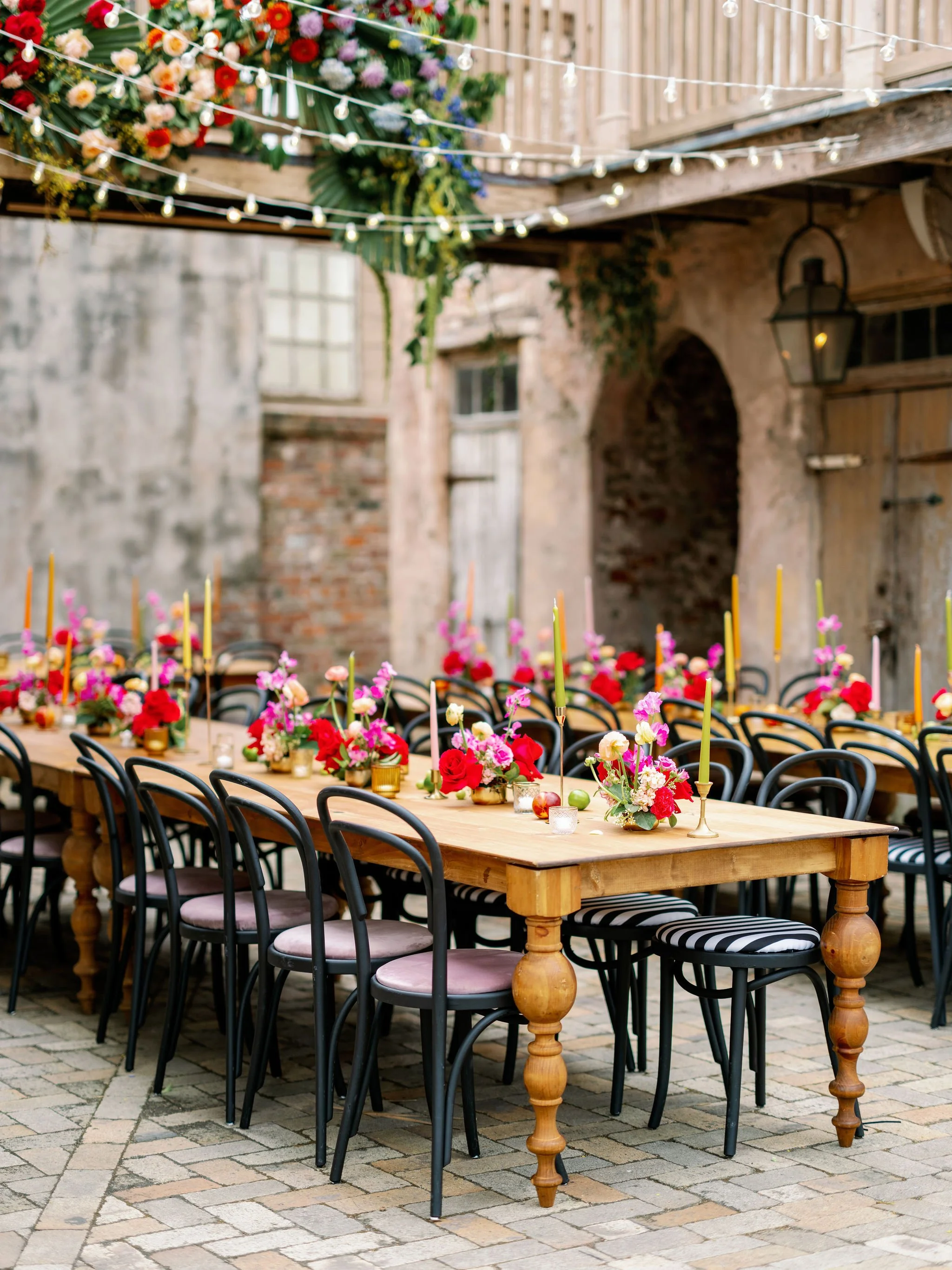 A long wooden table decorated with colorful flower arrangements, candles, and taper candles, set outdoors on cobblestone pavement, with black chairs around it and a rustic brick wall and lantern in the background.