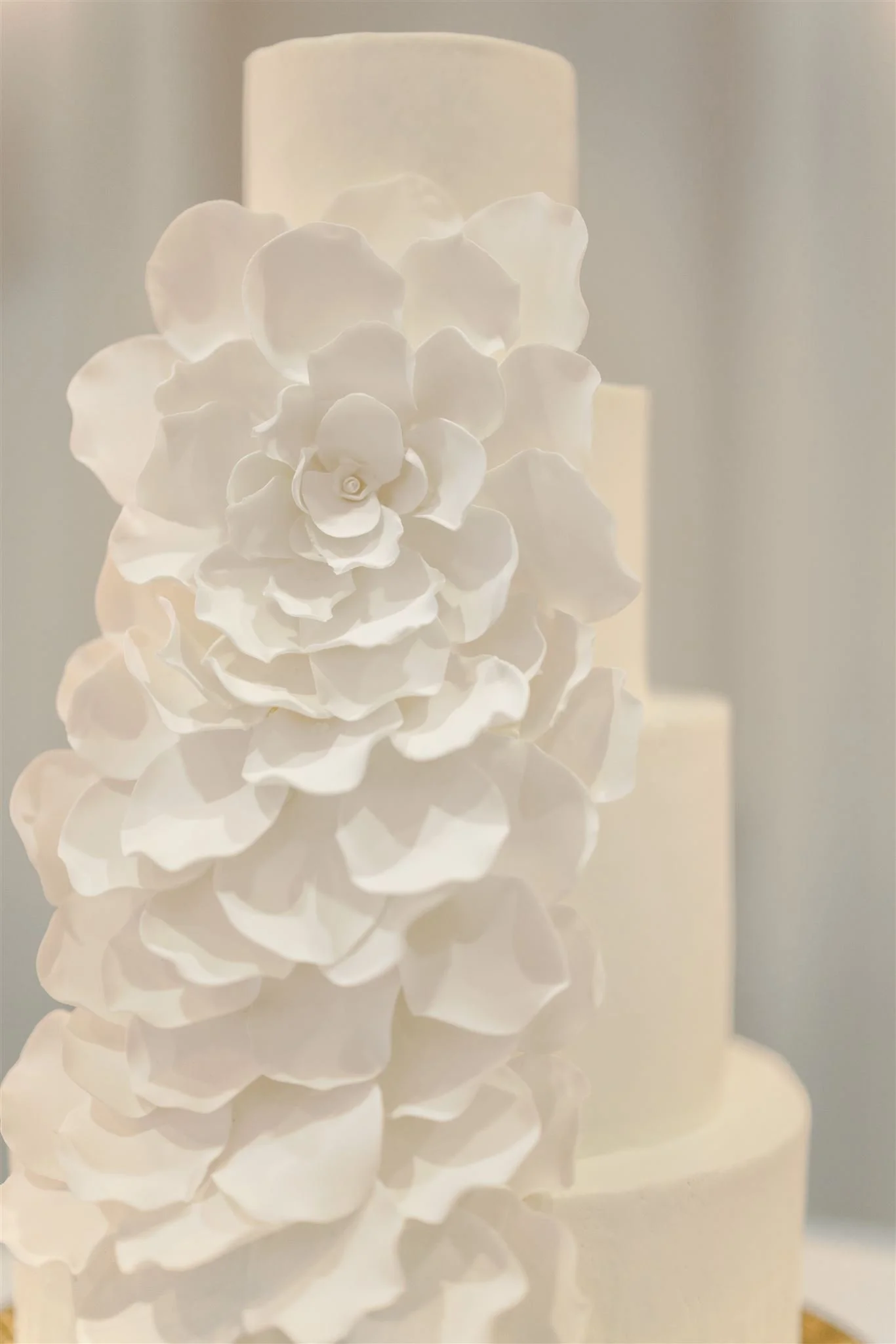 A white tiered cake decorated with white paper or fondant flowers, with a blurred background.