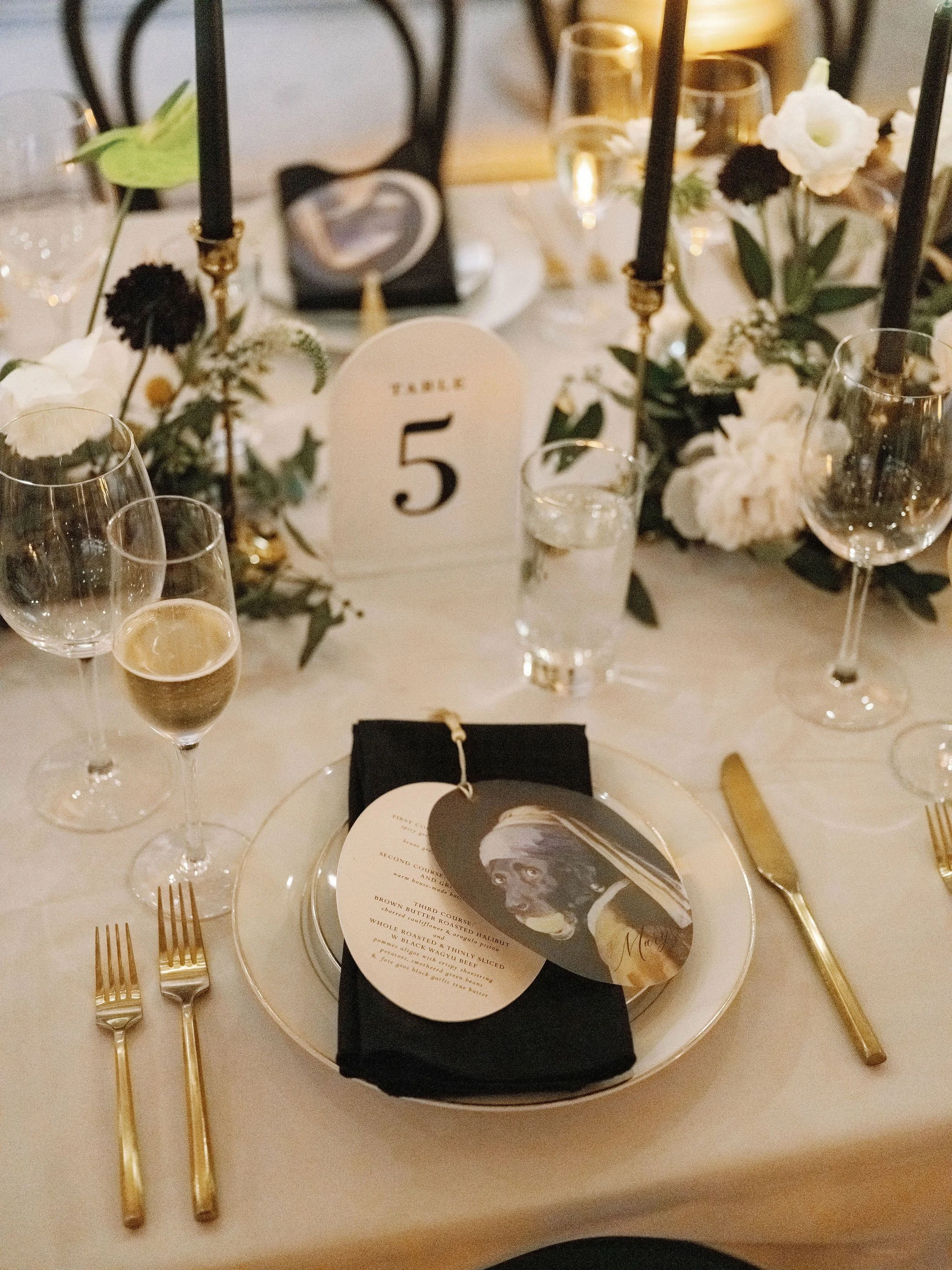 A table setting at a formal event with gold utensils, wine glasses, a menu, flowers, and a table number '5' card.