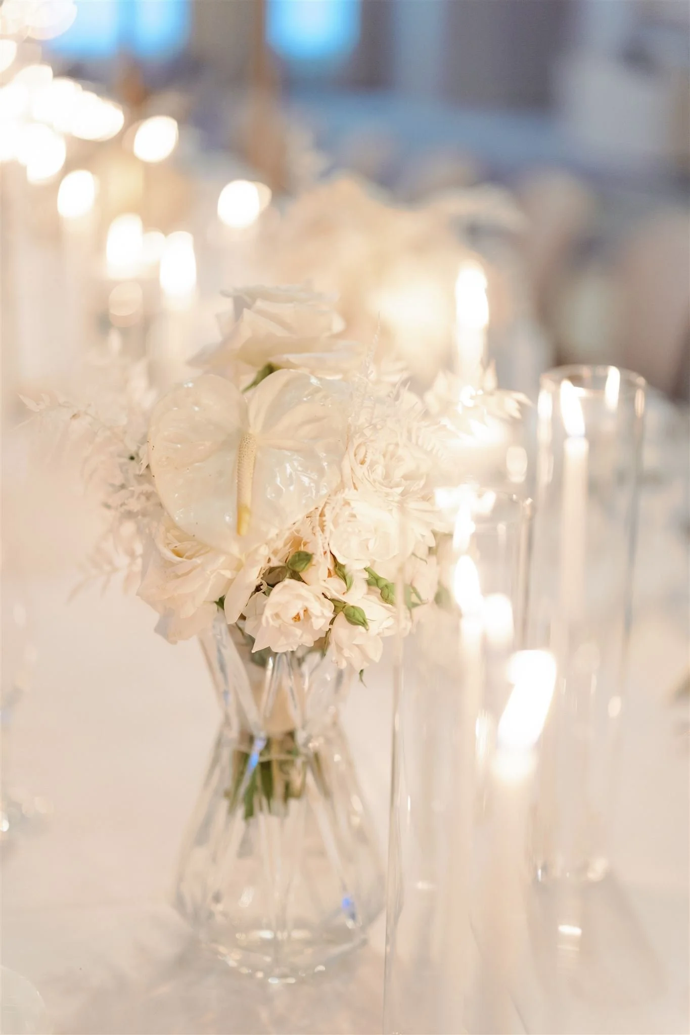 A bouquet of white flowers in a clear glass vase with multiple tall, slender glasses surrounding it, lit by warm, diffused lighting.