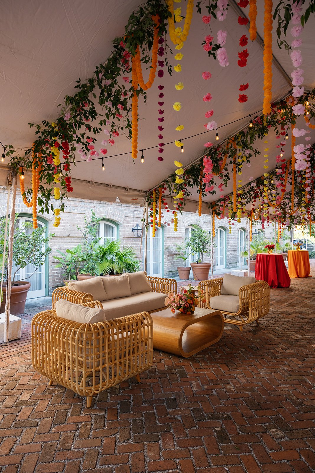 Courtyard reception at Hotel Peter and Paul in New Orleans with hanging flowers and string lights, wicker outdoor furniture, a wooden coffee table with flowers, and potted plants against brick building wall. Best New Orleans Indian wedding planner.