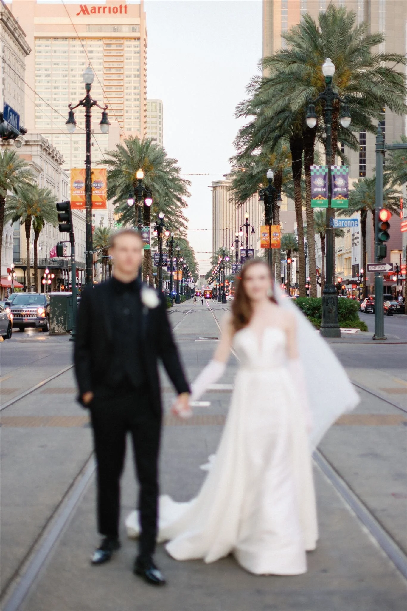 A bride and groom walking hand in hand down a city street lined with palm trees and streetlights, with tall buildings and banners in the background.