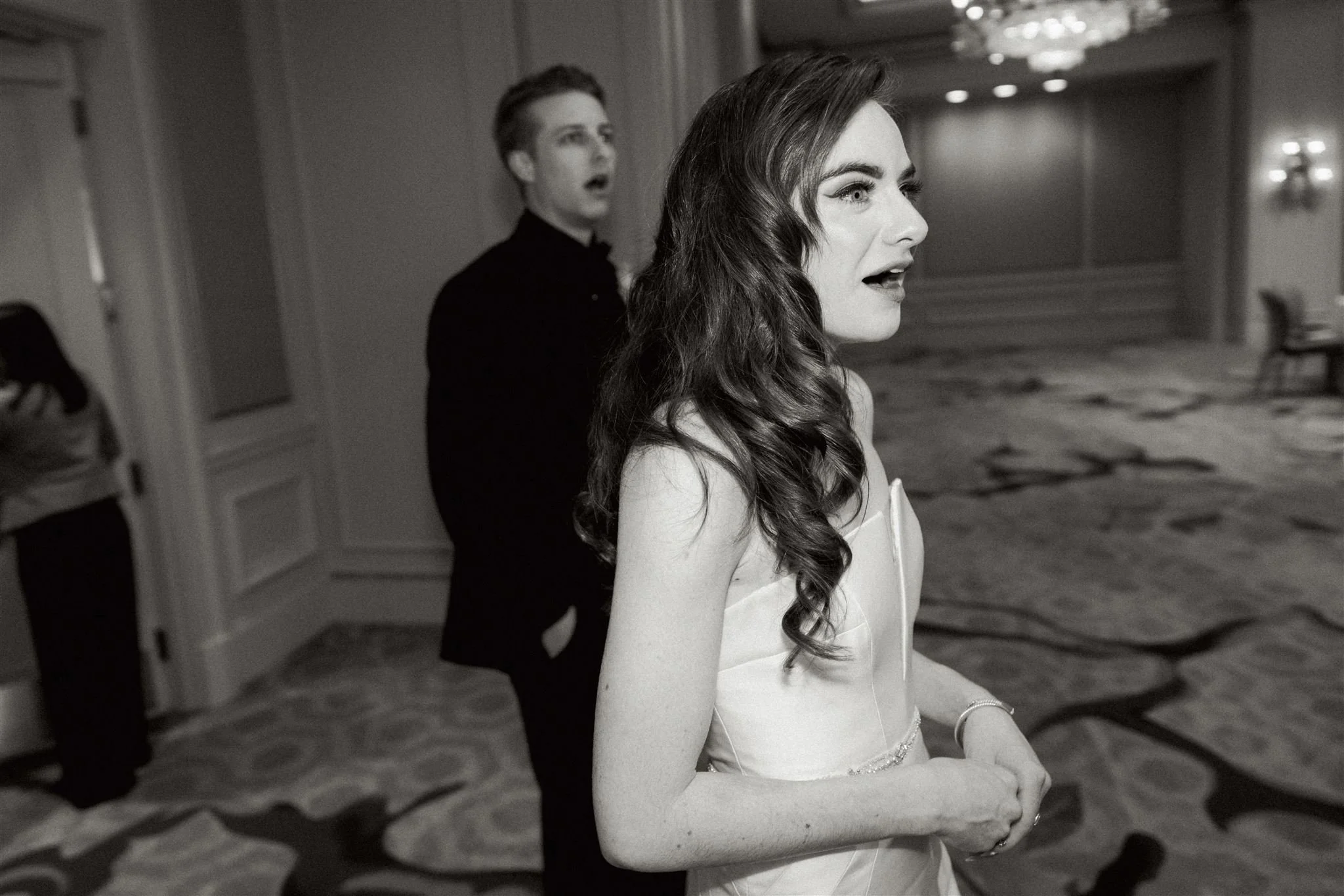Black and white photo of a woman with long, wavy hair and a strapless dress, possibly during a formal event or wedding, with a man in the background reacting with surprise or shock.