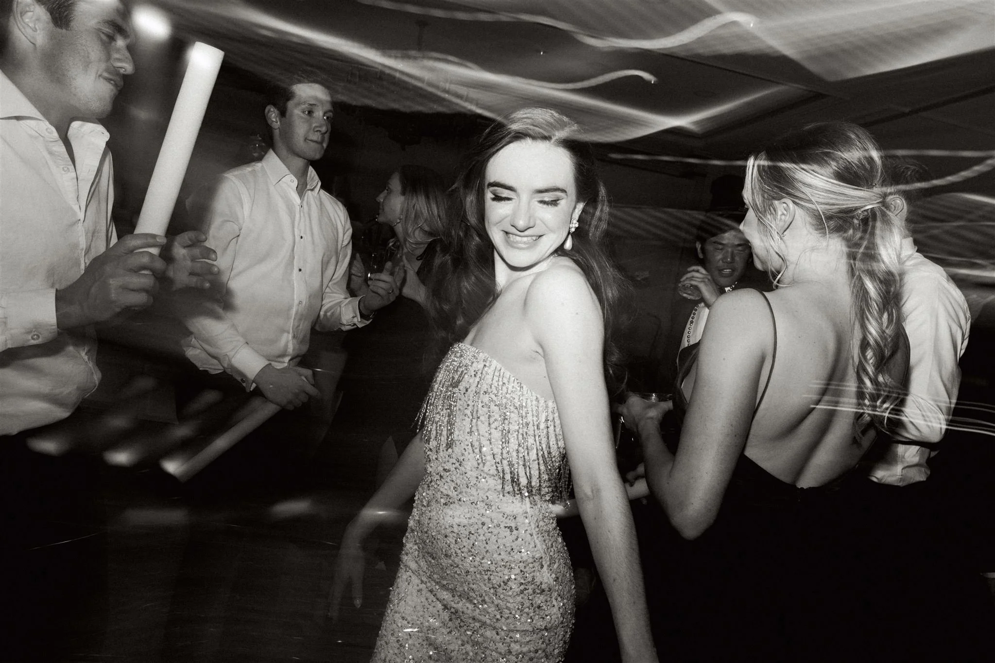 A woman in a shimmering gown with her eyes closed at a party or celebration, surrounded by other people, some holding drinks, in a lively indoor setting.