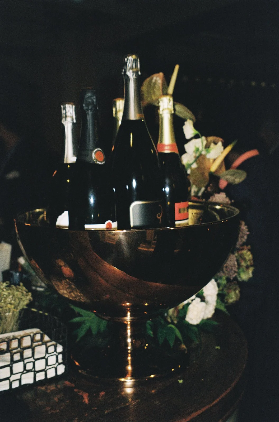 A metal bowl holding several bottles of champagne and a few cans, surrounded by flowers and greenery.