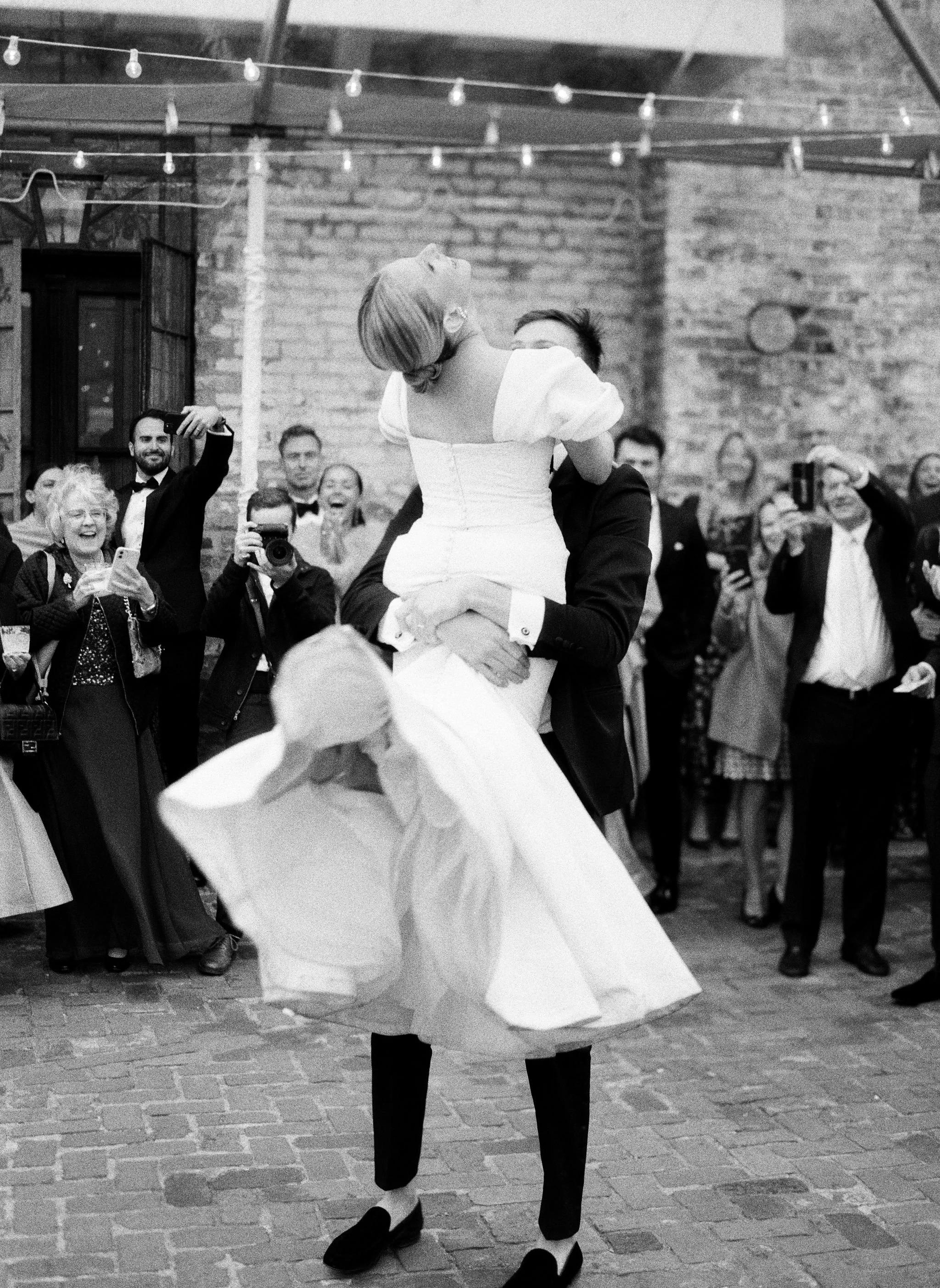 A person in a white dress with puffed sleeves is being lifted and spun around by someone in a dark suit at a celebration or wedding reception. Several guests are smiling, laughing, and taking photos in the background, with a brick wall and hanging st