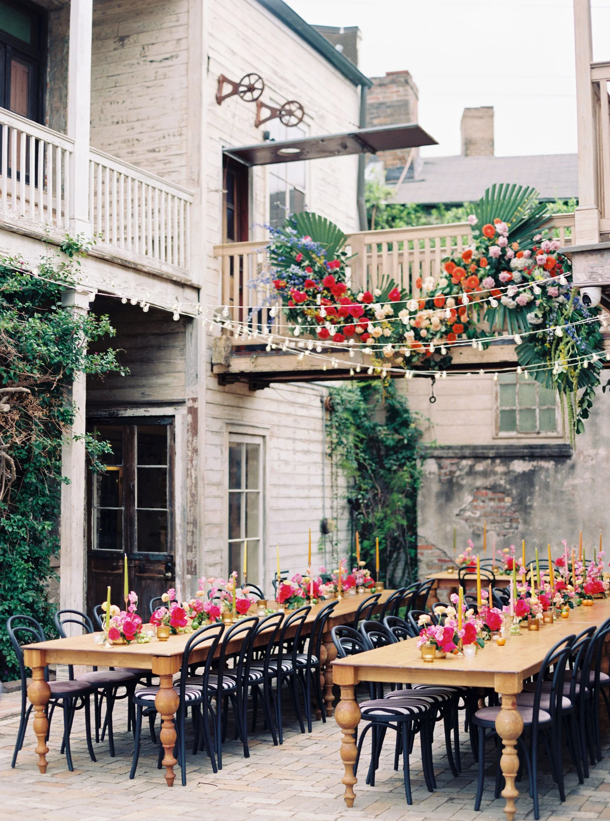 Outdoor wedding or event setup with long wooden tables decorated with pink flowers and candles, surrounded by black chairs with striped cushions, set against a rustic building with string lights and floral arrangements.
