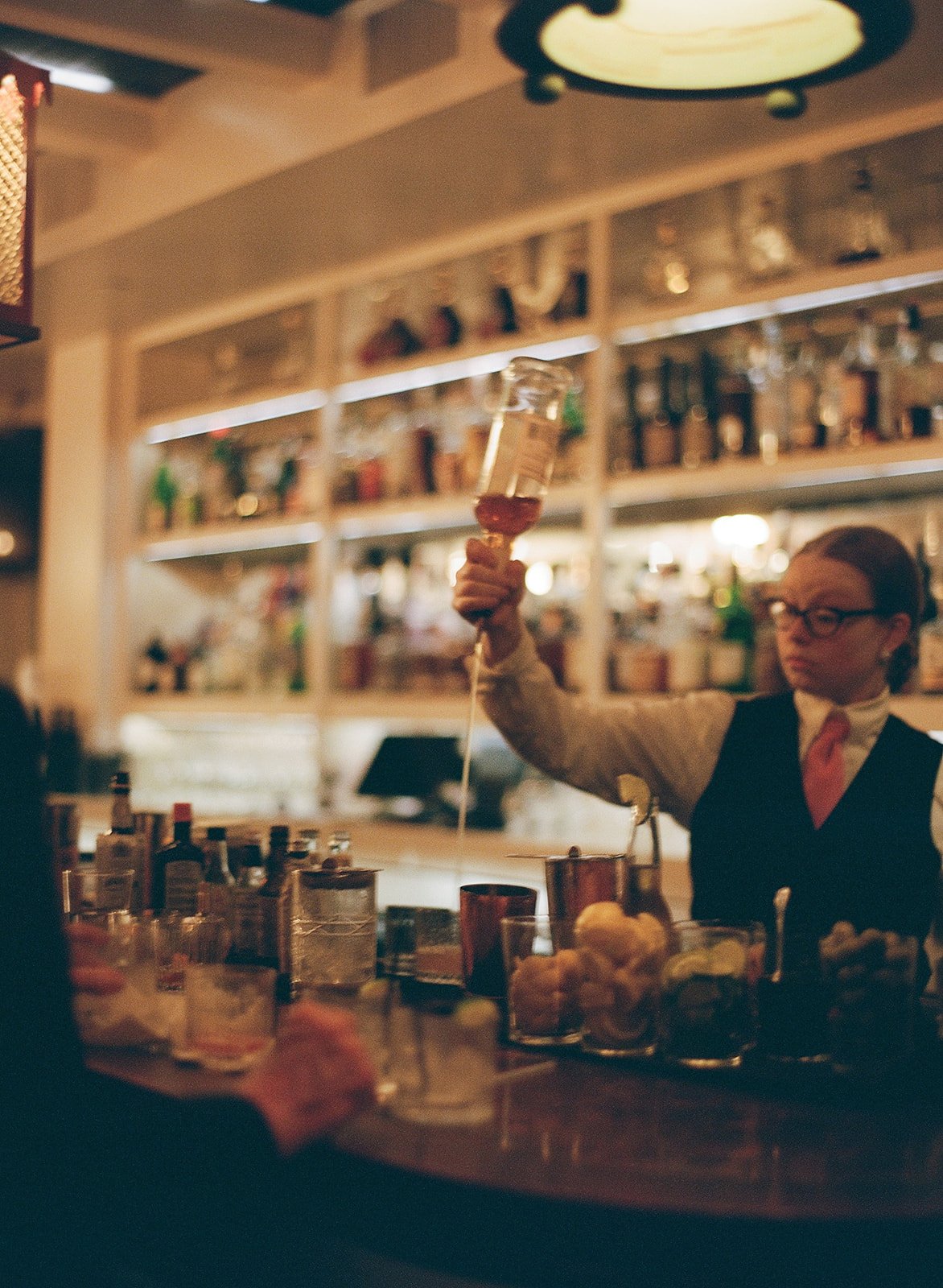Bartender pouring a cocktail at a bar with bottles on shelves in the background.