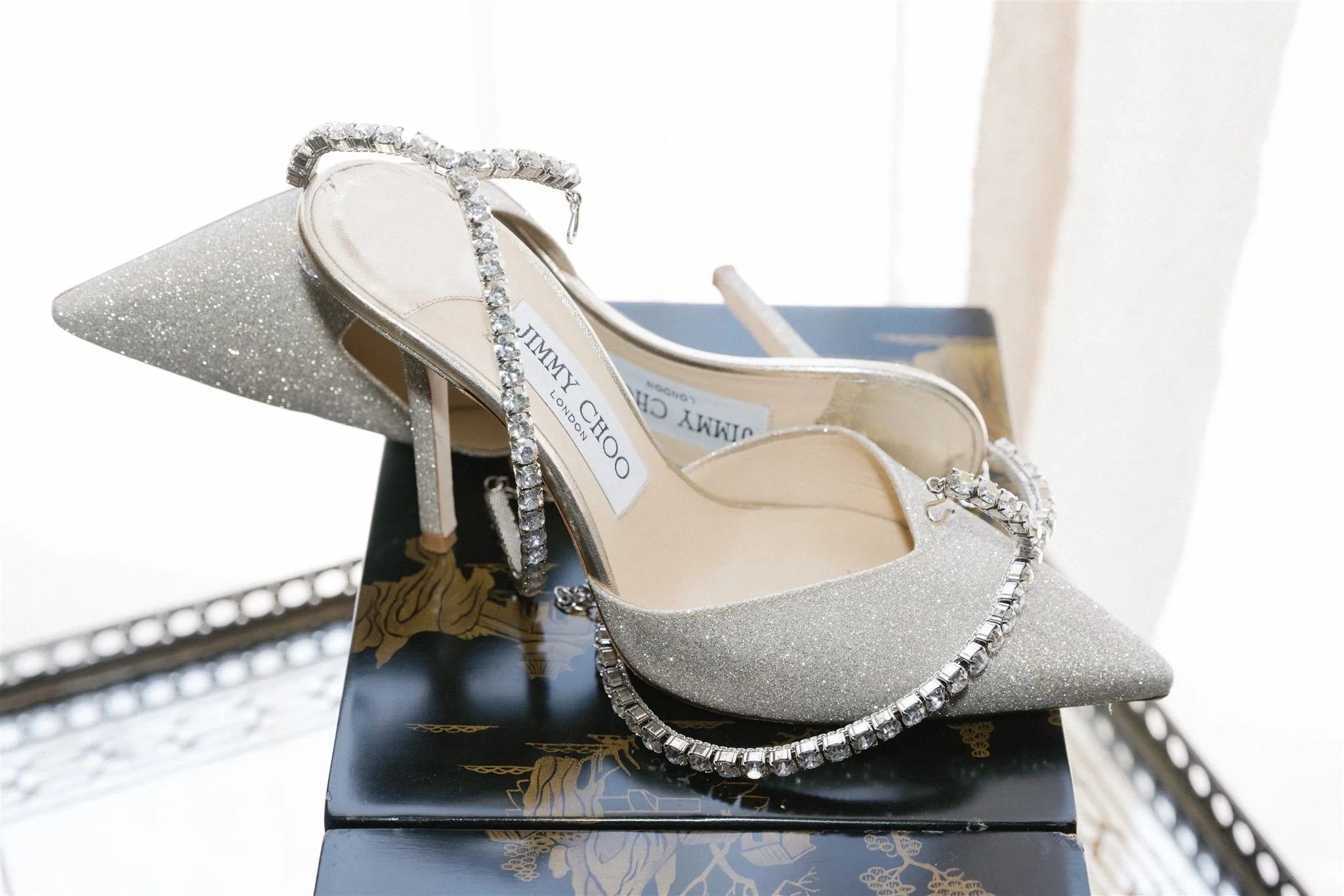 A pair of silver high-heeled shoes adorned with rhinestones, placed on a glittery shoe display block and a decorated black box.