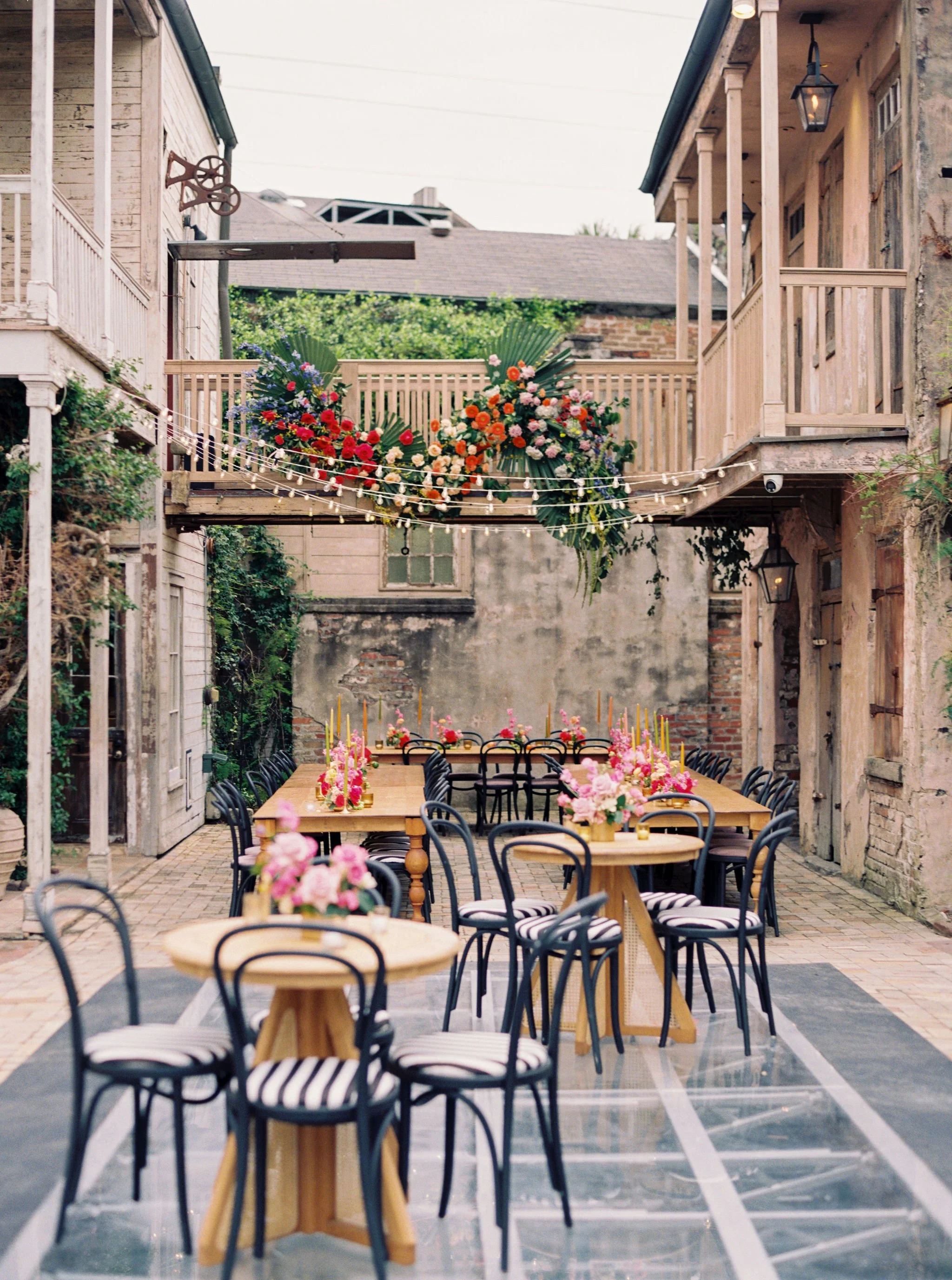 An outdoor wedding reception setup with long wooden tables and black chairs, decorated with pink floral centerpieces, in a courtyard with brick walls, string lights, and floral arrangements on a balcony.