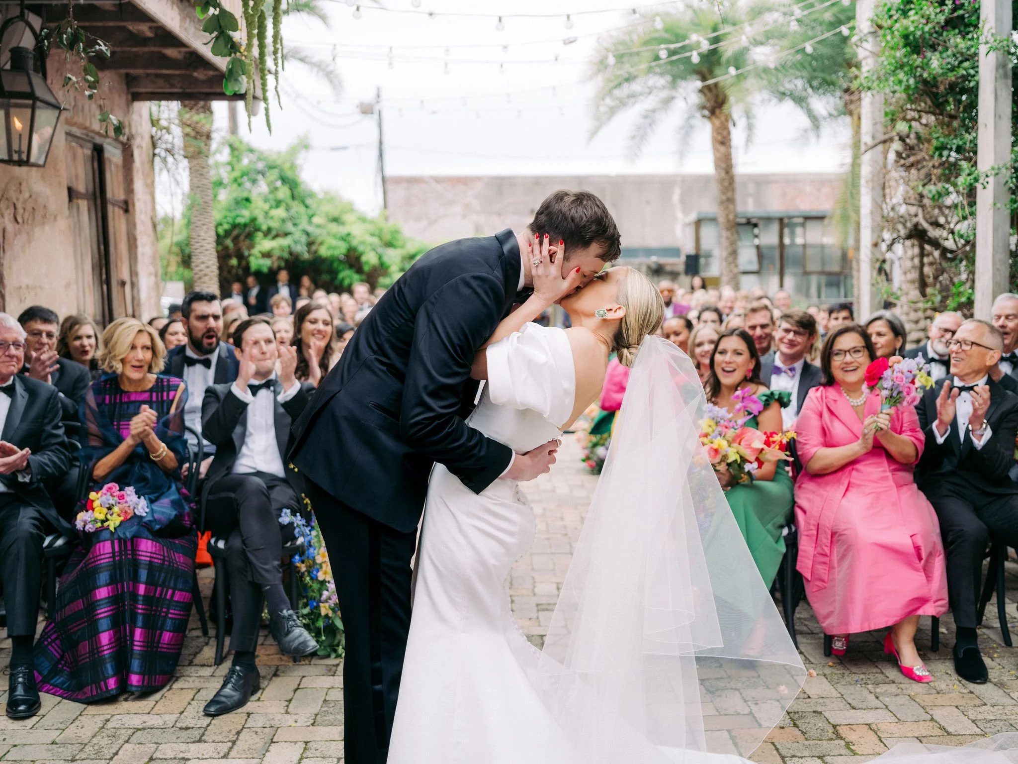 A Race and Religious courtyard wedding ceremony with a bride and groom kissing, surrounded by seated guests outdoors under string lights with trees and buildings in the background. Planned by the best wedding planner in New Orleans.