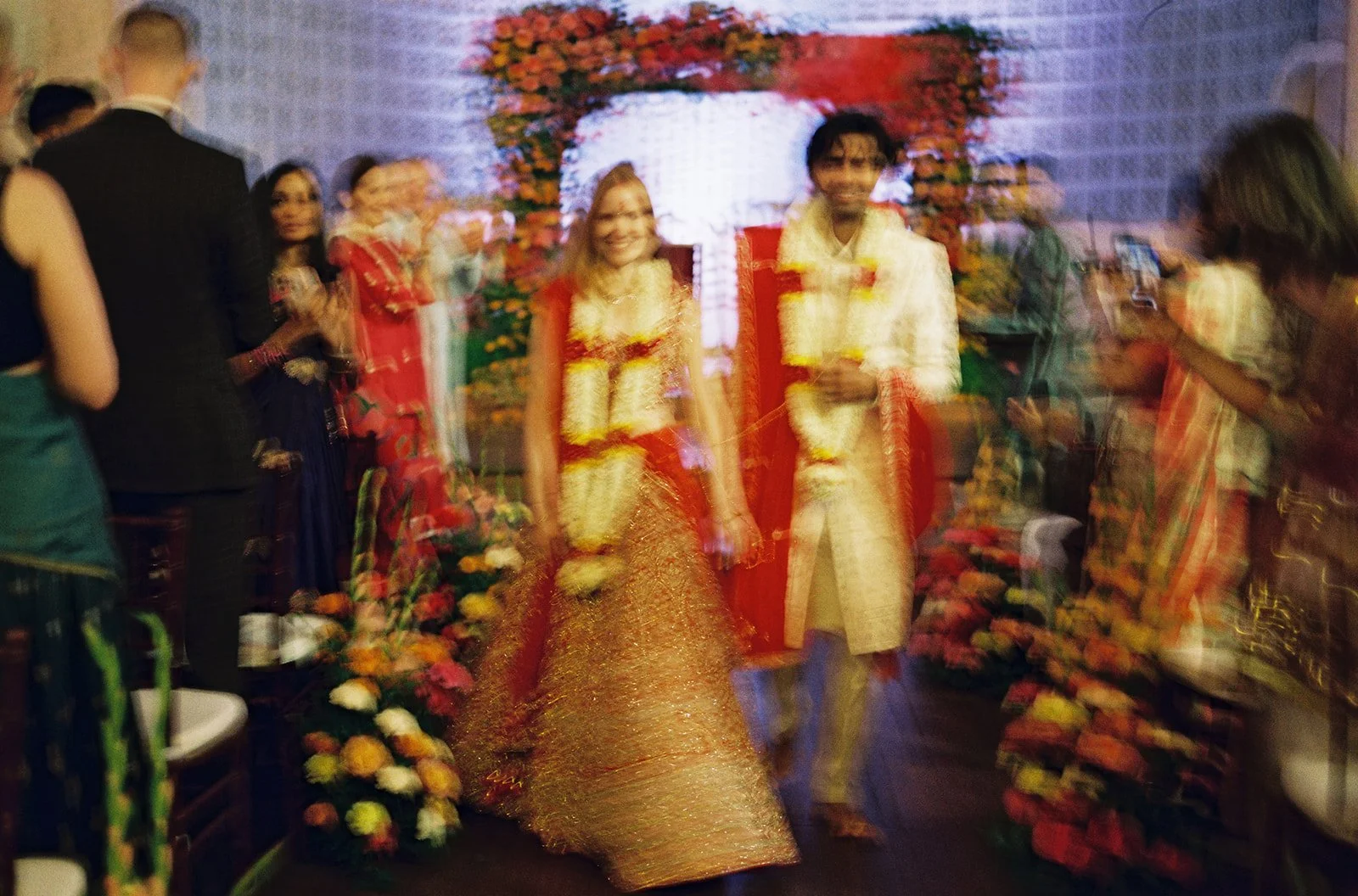 A couple dressed in traditional Indian wedding attire walking down a carpeted aisle, surrounded by guests and floral arrangements, at a wedding celebration.