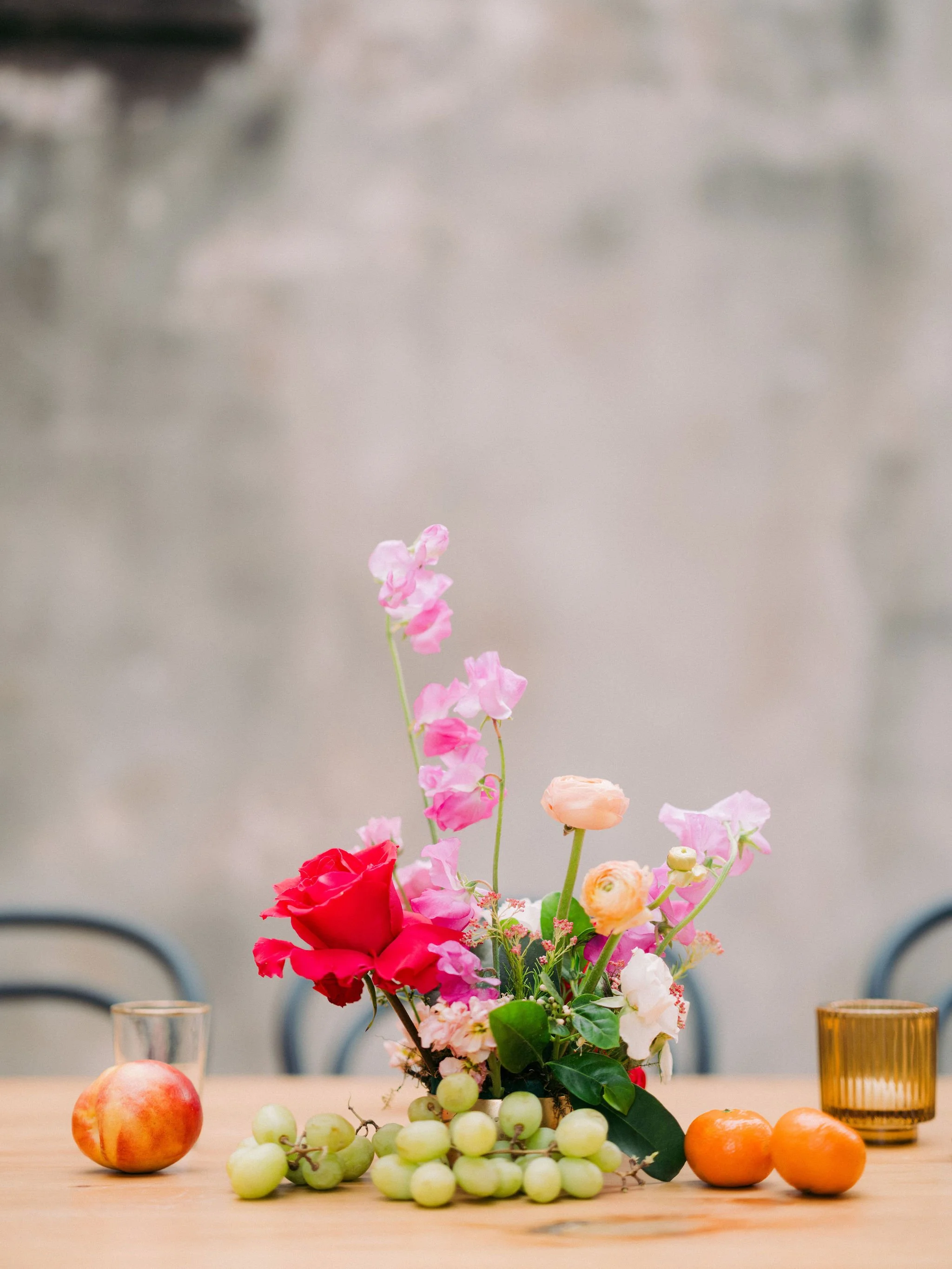 A floral centerpiece with pink, red, orange, and white flowers on a table, surrounded by apples, grapes, and oranges, with two glasses and a brown glass candle holder in the background.