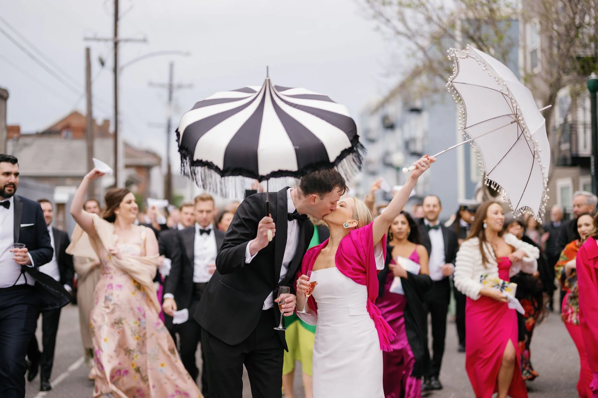 A joyful wedding second line in New Orleans with a bride and groom kissing under umbrellas, surrounded by guests in formal attire.