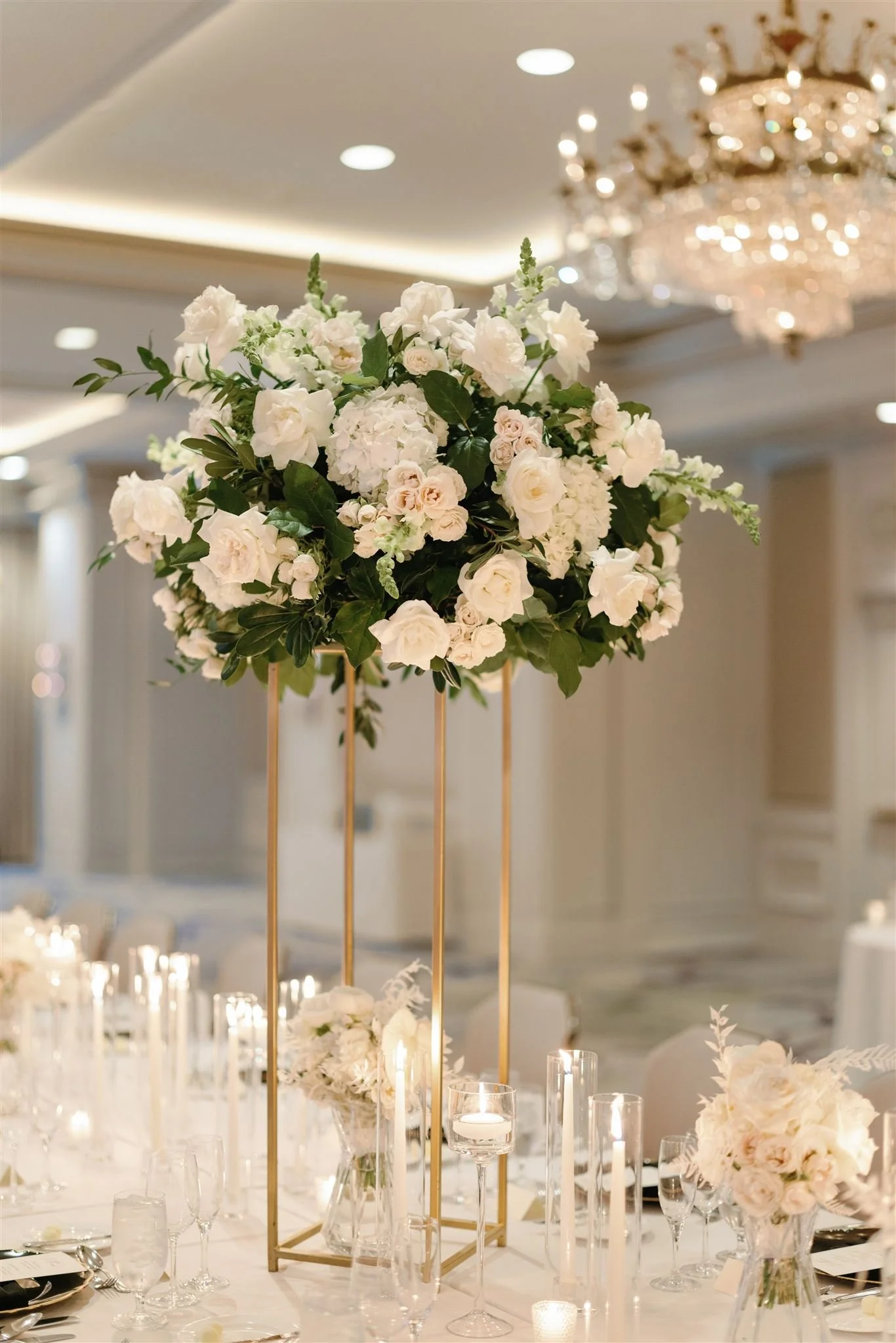 Elegant wedding reception table with tall floral centerpiece of white roses and greenery, surrounded by candles and glassware in a luxurious venue with chandelier lighting.