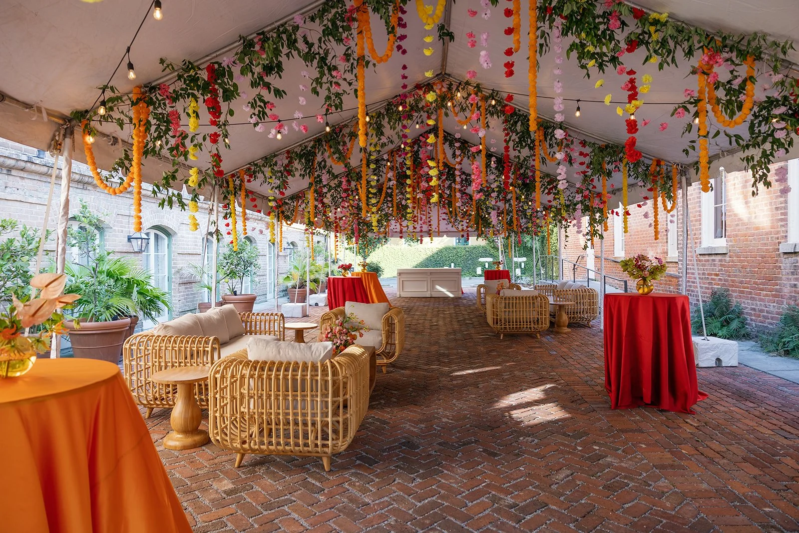 Courtyard reception at Hotel Peter and Paul in New Orleans with hanging flower garlands, string lights, wicker furniture, round tables with orange tablecloths, and potted plants along brick walls. Best Indian wedding planner in New Orleans.