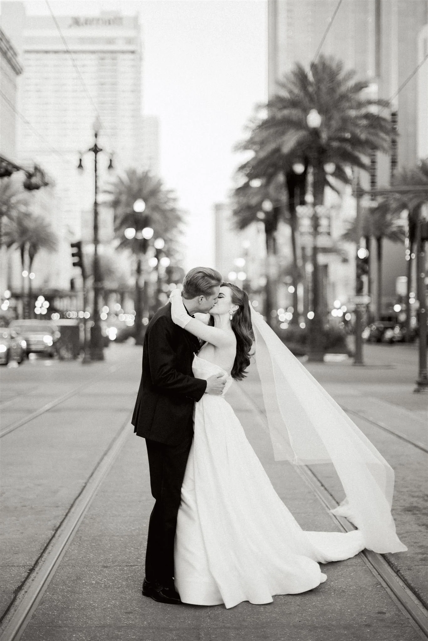 A newlywed couple sharing a kiss on a city street with palm trees and tall buildings in the background, black and white photo.