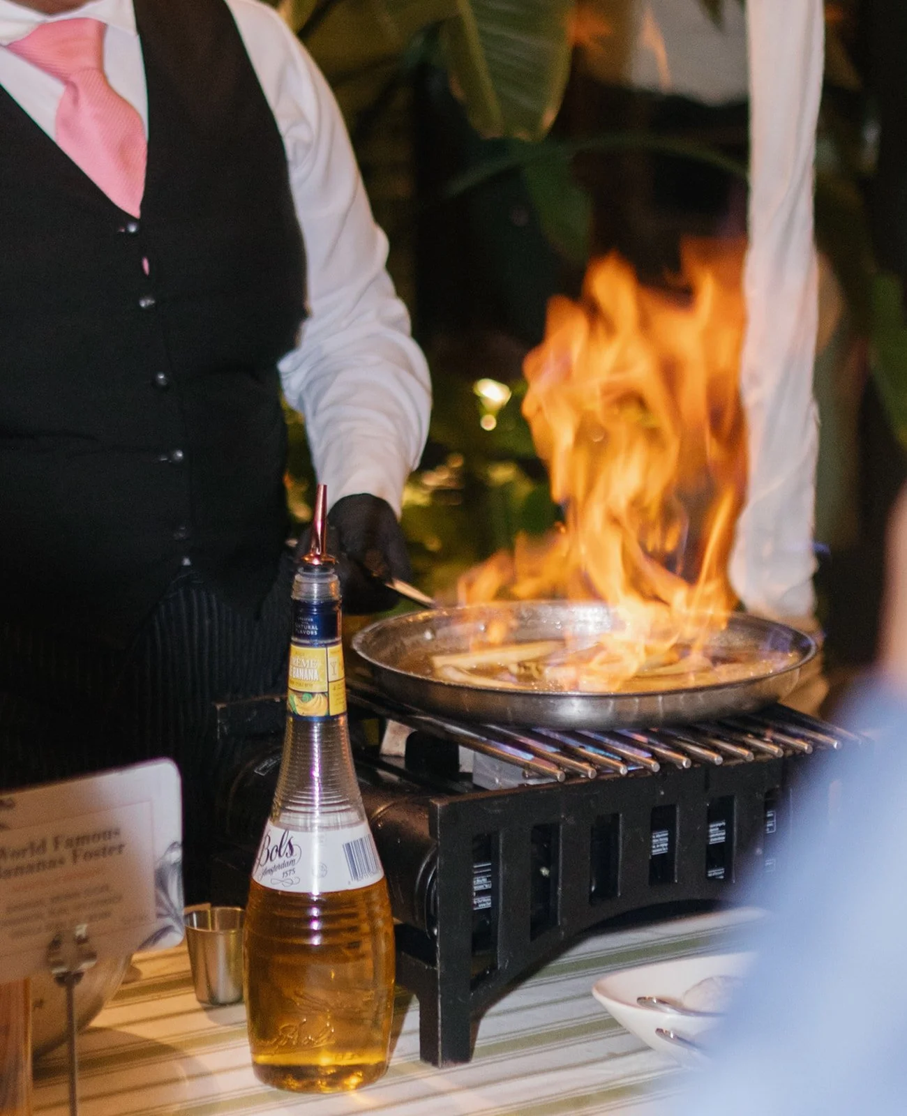 Chef cooking with flames in a pan at a restaurant or outdoor setting, with a bottle of syrup and other table items visible in the foreground.
