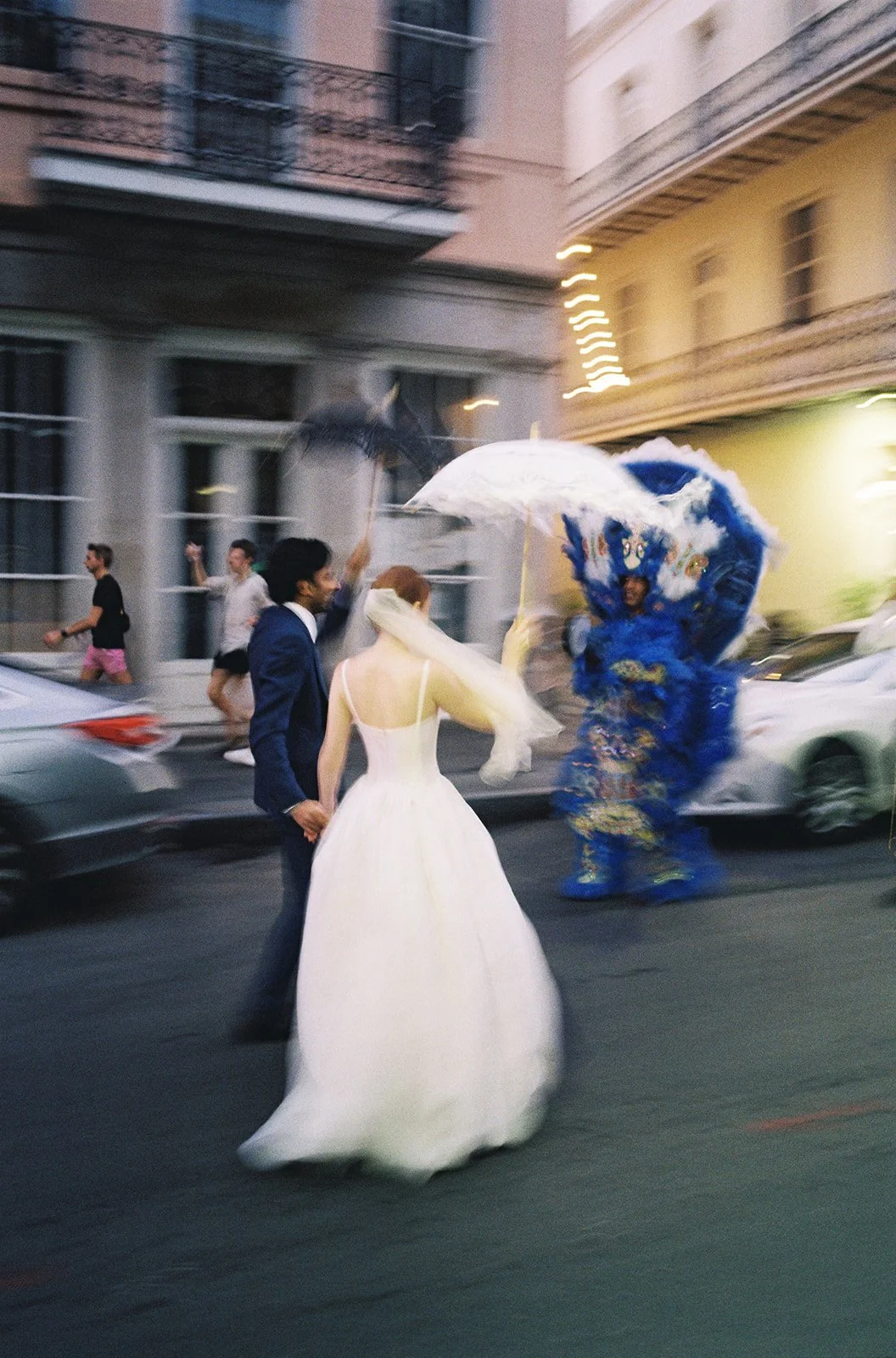 Couple in wedding attire standing on a city street, with a performer in elaborate costume nearby, holding umbrellas and people walking in the background. New Orleans wedding second line with Mardi Gras Indians.